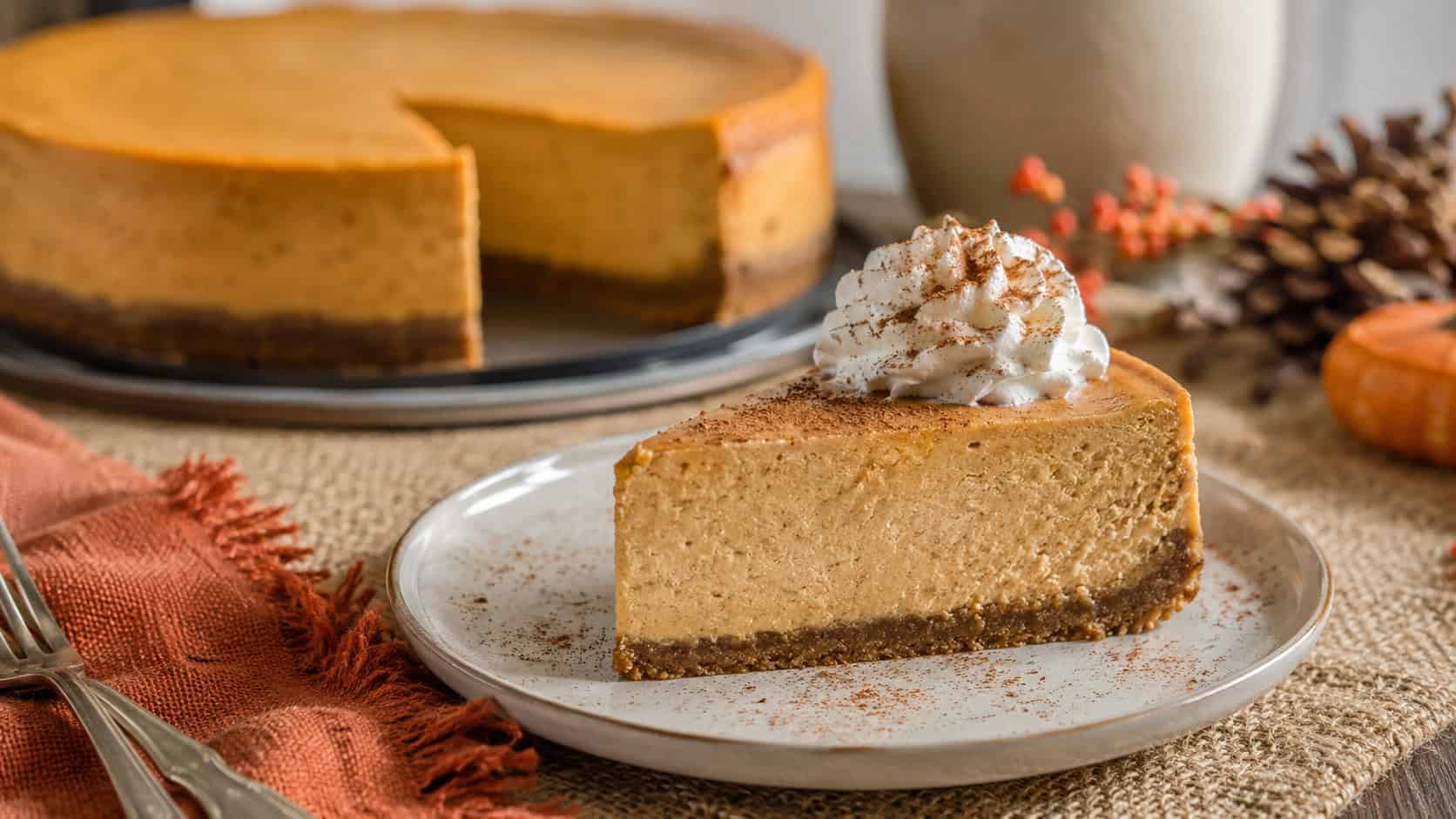 a creamy pumpkin spice cheesecake slice on a small plate with a dollop of whipped cream and a dusting of cinnamon in a wide horizontal shot, whole cheesecake softly blurred behind, festive table setting, natural light, shallow depth of field, landscape orientation