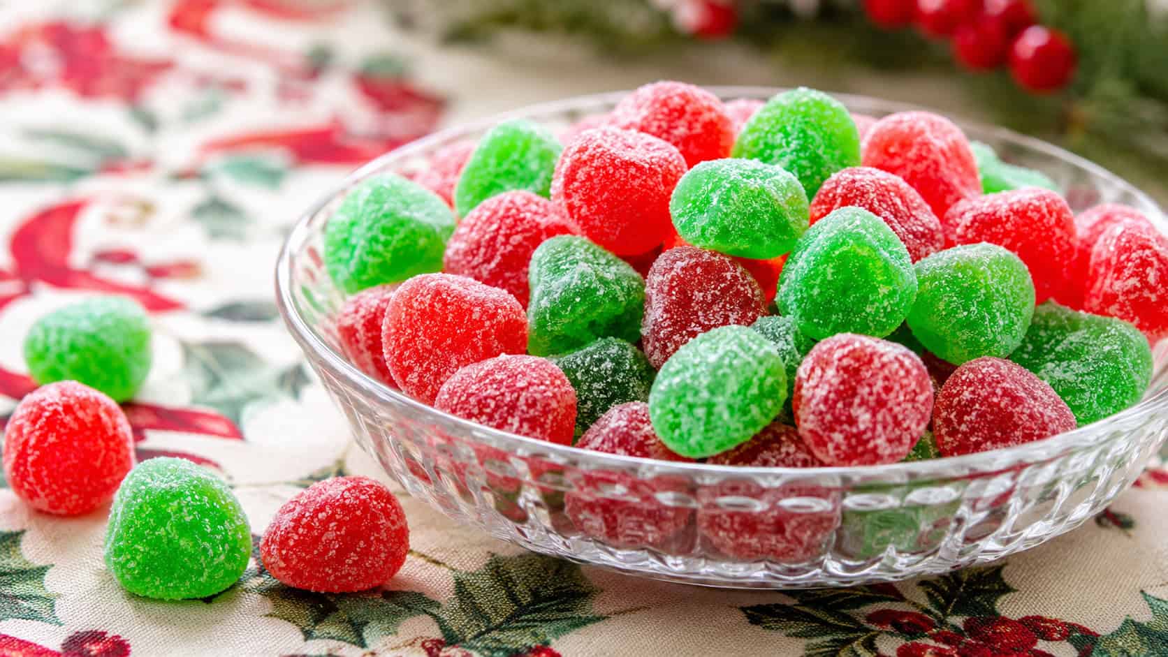 bright red and green sugar-coated gumdrops heaped in a glass dish in a wide horizontal shot, a holiday tablecloth and scattered candies beside it, soft bright light, shallow depth of field, landscape orientation