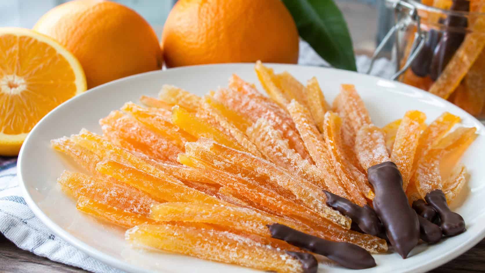 sugar-coated candied orange peel strips fanned on a white plate in a wide horizontal shot, fresh oranges and a few chocolate-dipped pieces nearby, bright natural light, shallow depth of field, landscape orientation