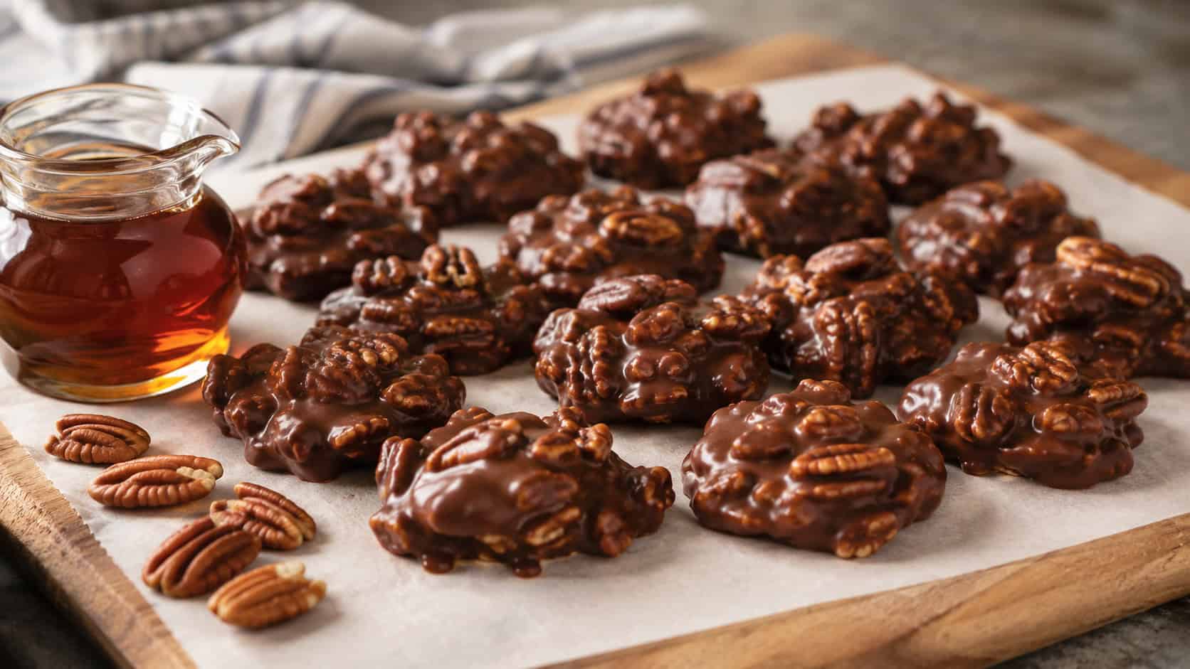glossy chocolate pecan clusters on a parchment-lined board in a wide horizontal shot, a small jug of maple syrup and loose pecans nearby, warm natural light, shallow depth of field, landscape orientation