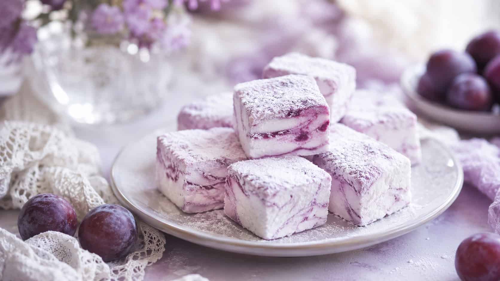 soft purple-swirled marshmallow squares dusted with powdered sugar on a pale plate in a wide horizontal shot, a lace cloth and sugar plums nearby, soft dreamy light, shallow depth of field, landscape orientation