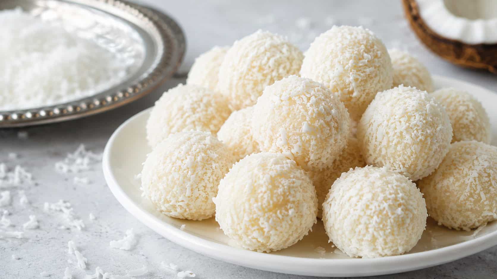 round coconut snowball truffles coated in shredded coconut piled on a white plate in a wide horizontal shot, scattered coconut and a silver tray nearby, soft bright light, shallow depth of field, landscape orientation