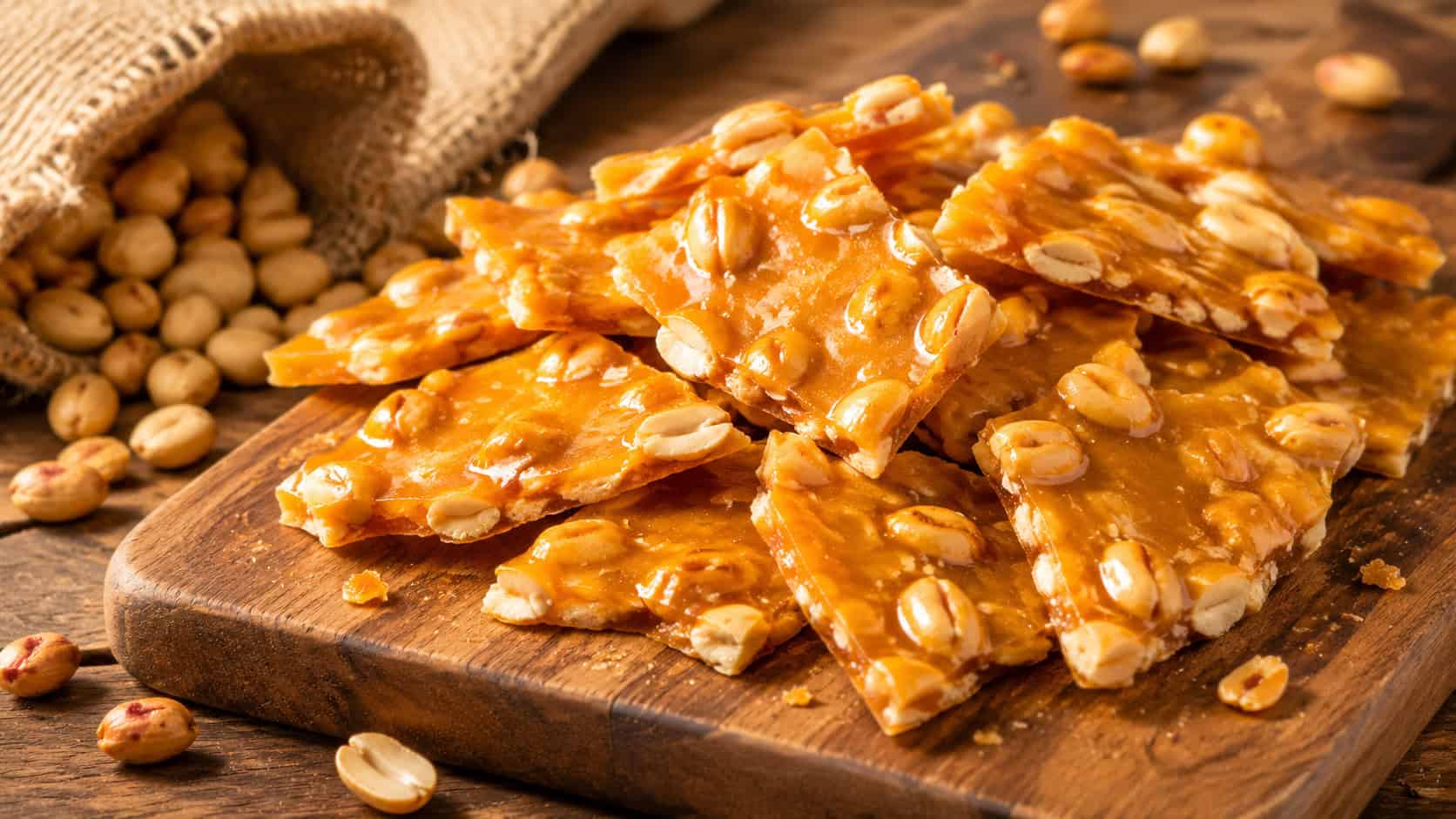 broken shards of golden peanut brittle piled on a wooden board in a wide horizontal shot, loose roasted peanuts and a burlap cloth nearby, warm natural light, shallow depth of field, landscape orientation