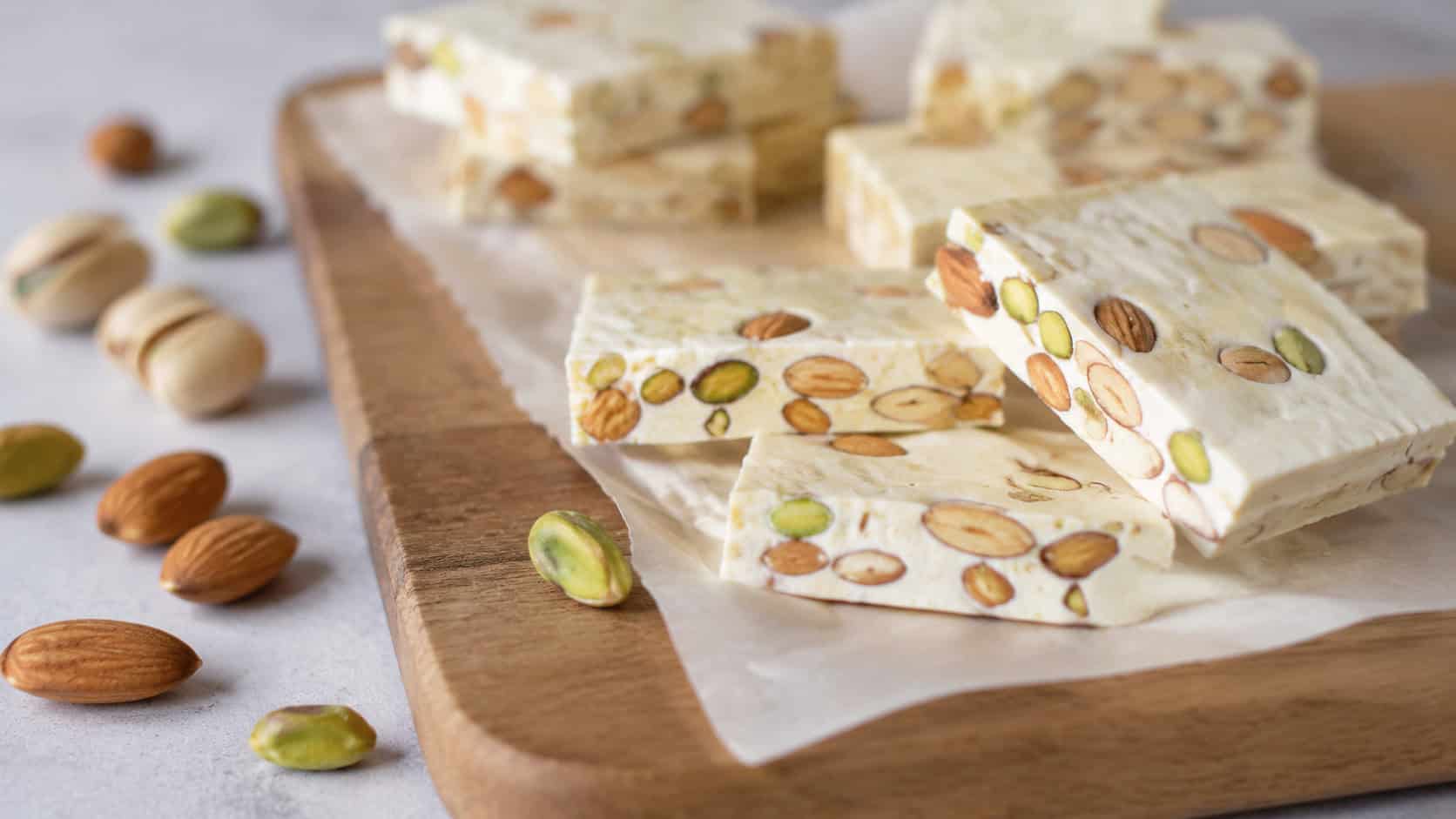 pale chewy nougat bars studded with whole almonds and green pistachios on parchment in a wide horizontal shot, a wooden board and scattered nuts beside them, soft natural light, shallow depth of field, landscape orientation
