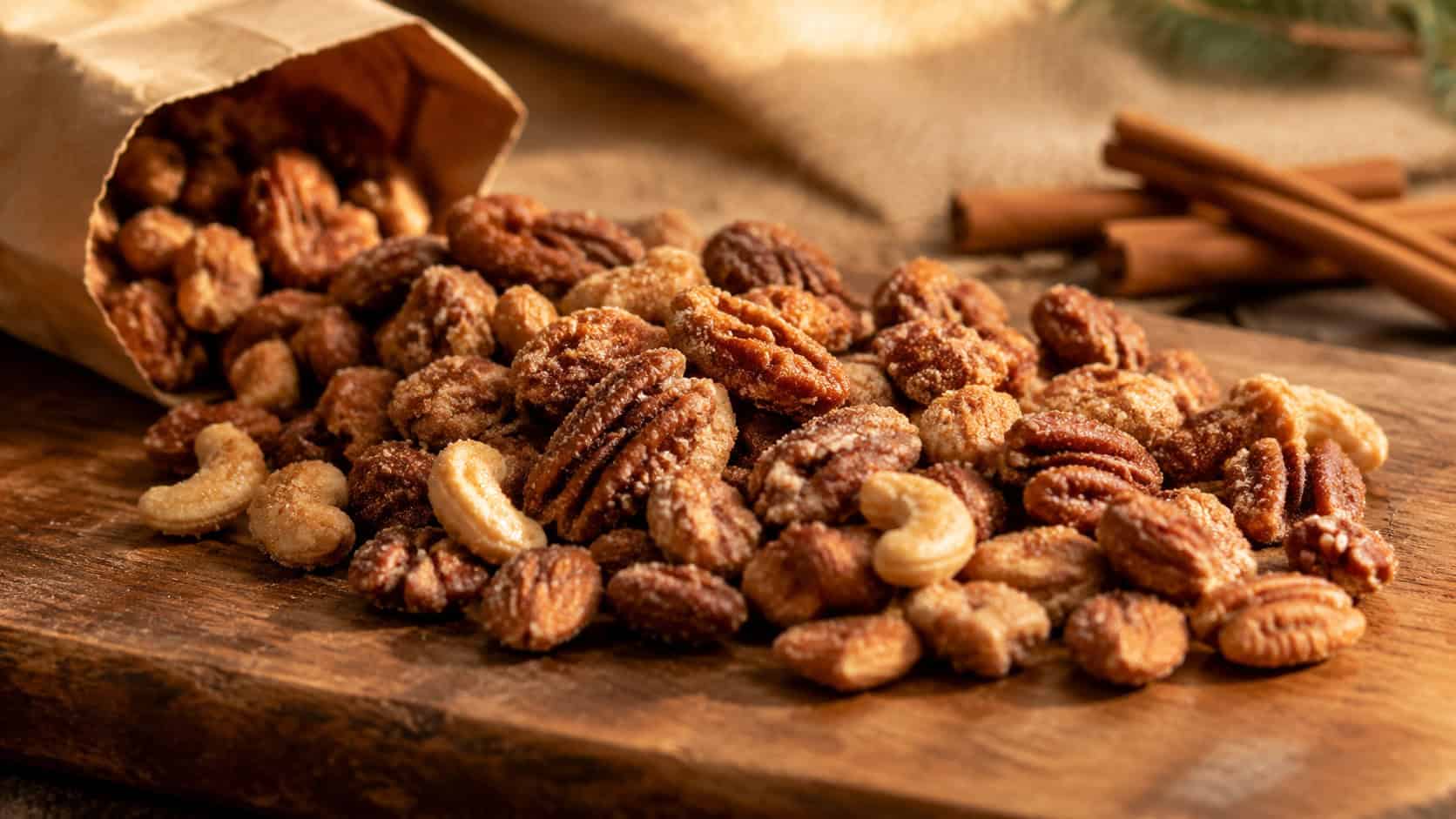 glistening cinnamon-sugar coated mixed nuts spread on a rustic wooden board in a wide horizontal shot, a kraft paper bag and cinnamon sticks nearby, warm golden light, shallow depth of field, landscape orientation