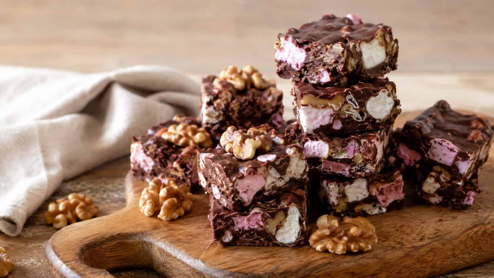 chunky rocky road fudge squares packed with marshmallows and walnuts on a wooden board in a wide horizontal shot, loose walnuts and a linen cloth nearby, warm natural light, shallow depth of field, landscape orientation