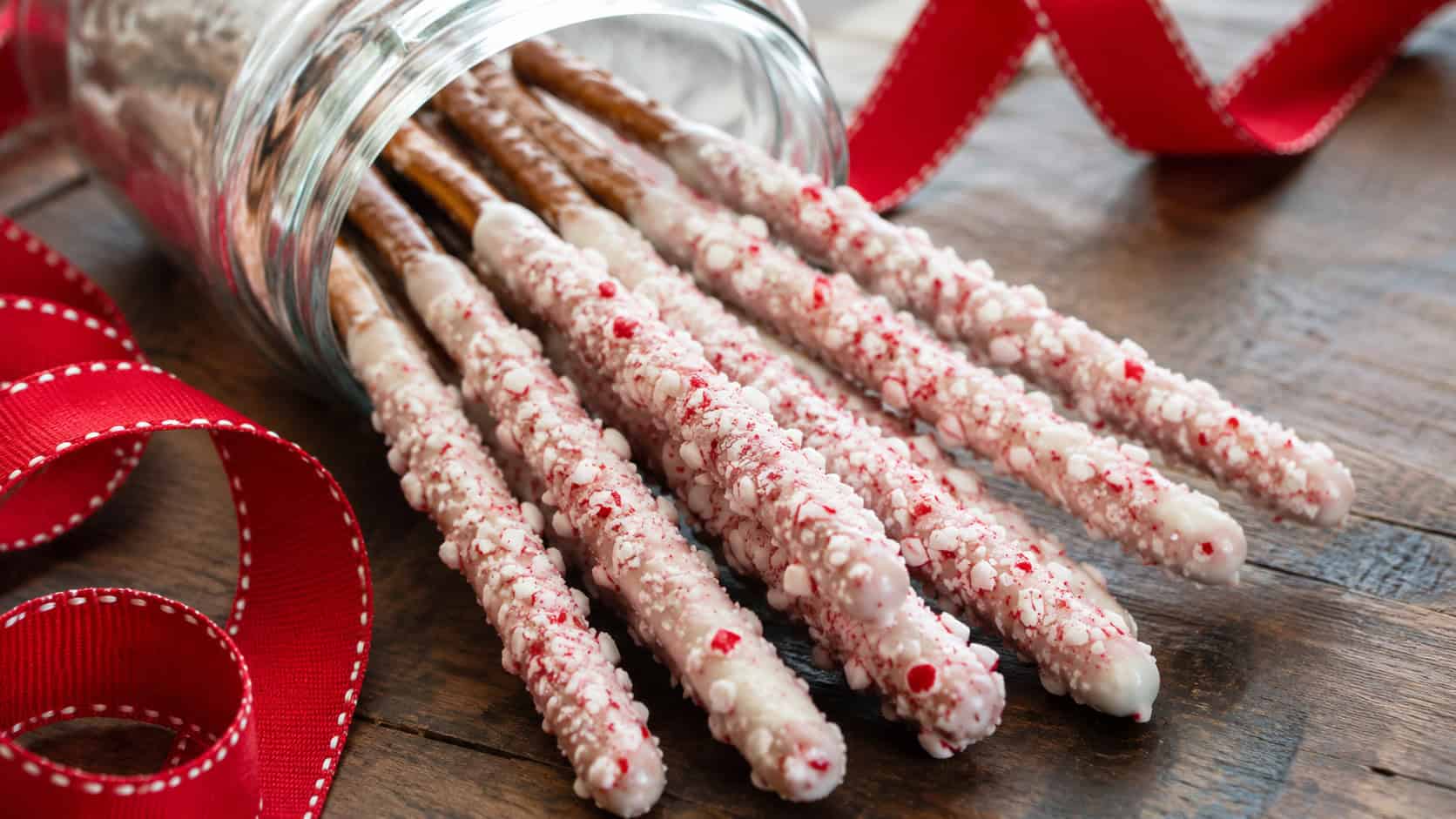 white-chocolate-dipped pretzel rods rolled in crushed peppermint candy, arranged in a wide horizontal row in a tall glass jar tipped on a wooden table, festive red ribbon nearby, bright natural light, shallow depth of field, landscape orientation