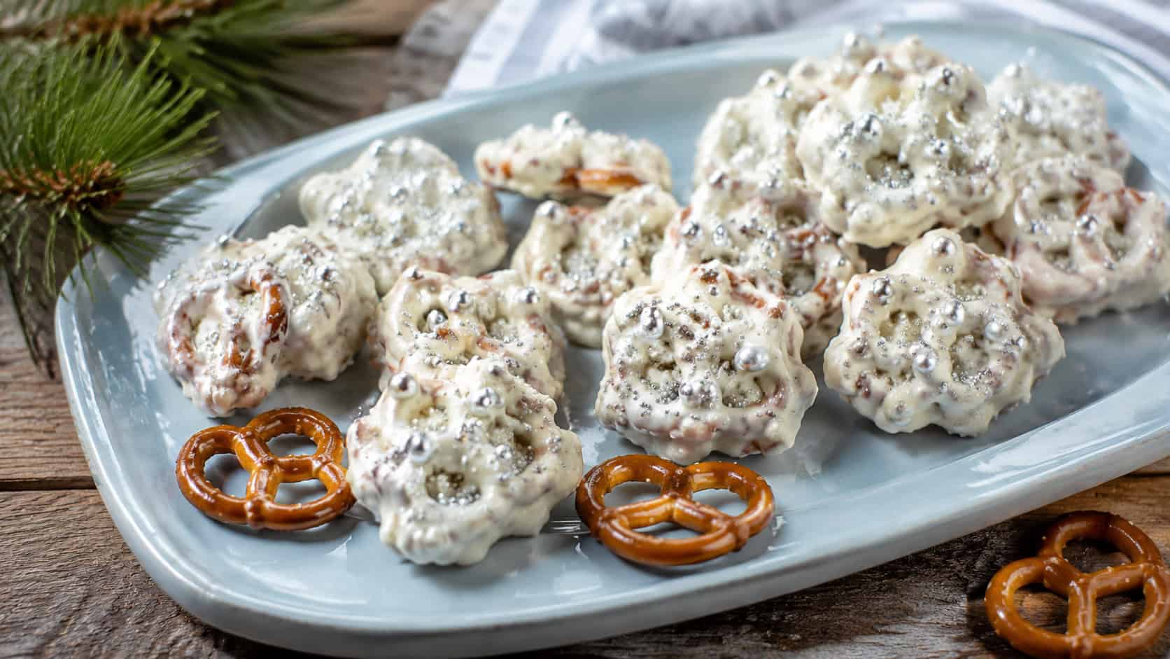 white chocolate pretzel clusters topped with silver pearls scattered across a pale blue ceramic platter in a wide horizontal shot, a few loose pretzels and a sprig of pine alongside, crisp natural light, shallow depth of field, landscape orientation