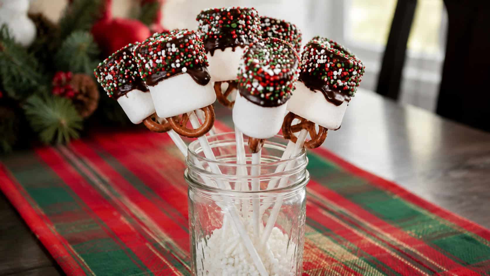 marshmallow and pretzel pops on sticks standing in a tall mason jar in a wide horizontal shot, tops dipped in chocolate and red and green sprinkles, festive table runner underneath, bright cheerful natural light, shallow depth of field, landscape orientation