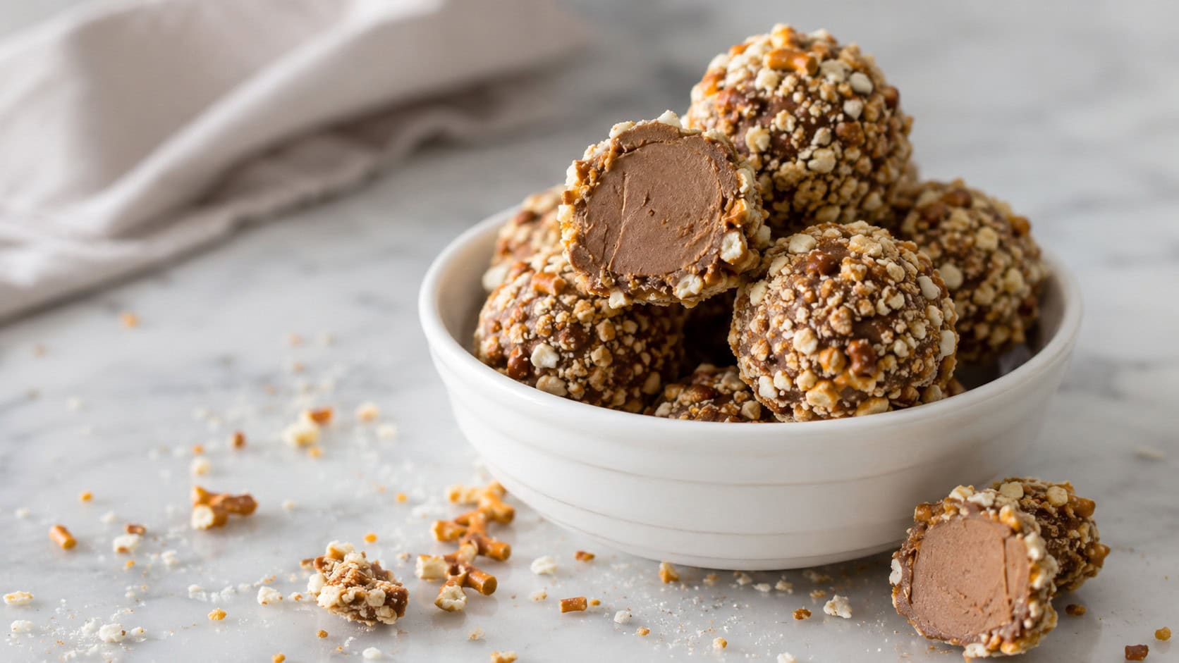 hazelnut truffles rolled in crushed pretzel coating piled in a small white bowl in a wide horizontal shot, a few cut open to show the creamy center, dusting of crumbs on a marble surface, soft natural light, shallow depth of field, landscape orientation