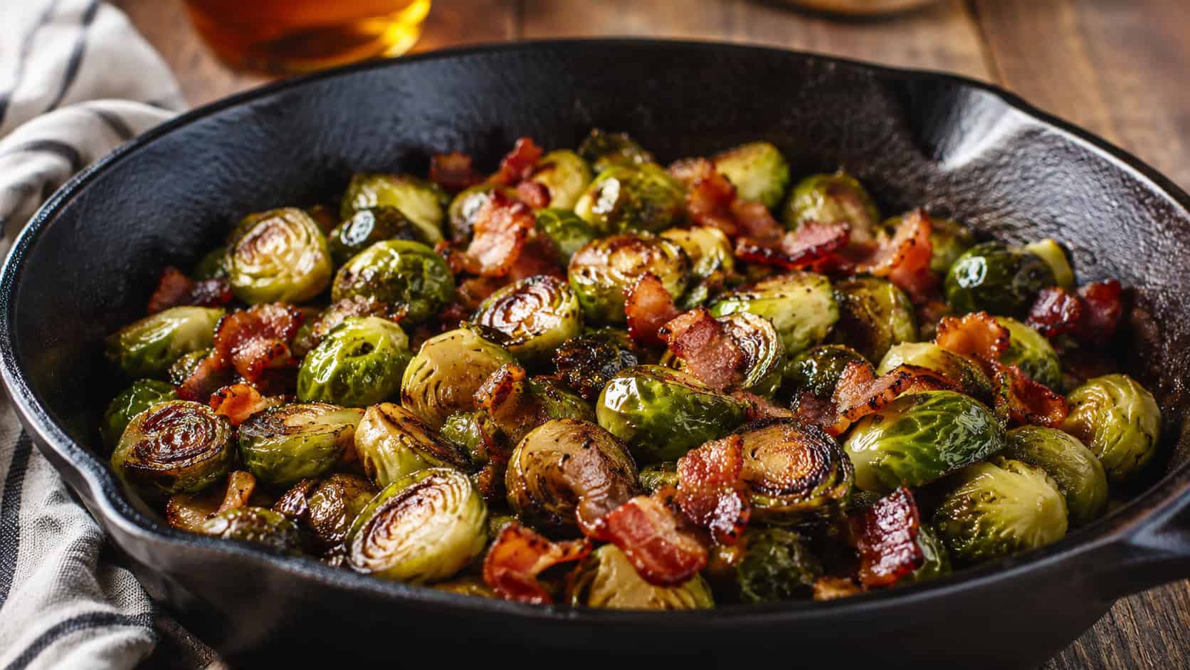 caramelized roasted brussels sprouts tossed with crispy bacon and glistening maple glaze, served in a wide horizontal composition in a cast iron skillet on a rustic table, warm natural light, shallow depth of field, landscape orientation
