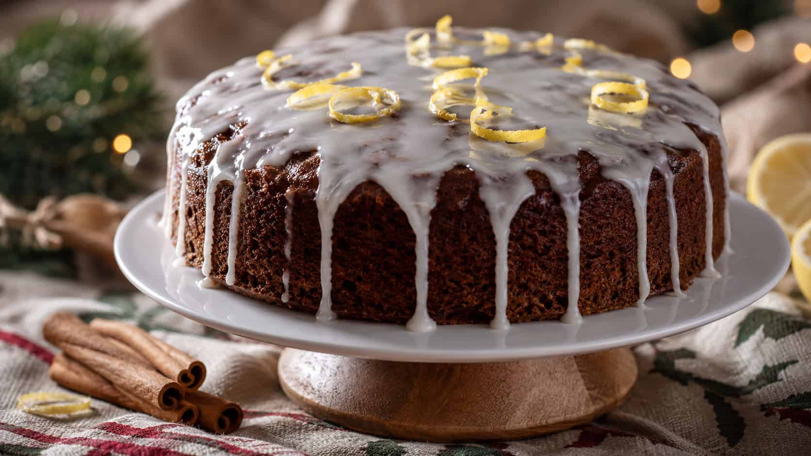 a dark spiced gingerbread cake drizzled with white lemon glaze and a few lemon curls, shown in a wide horizontal composition on a cake stand over a festive linen with cinnamon sticks, warm natural light, shallow depth of field, landscape orientation