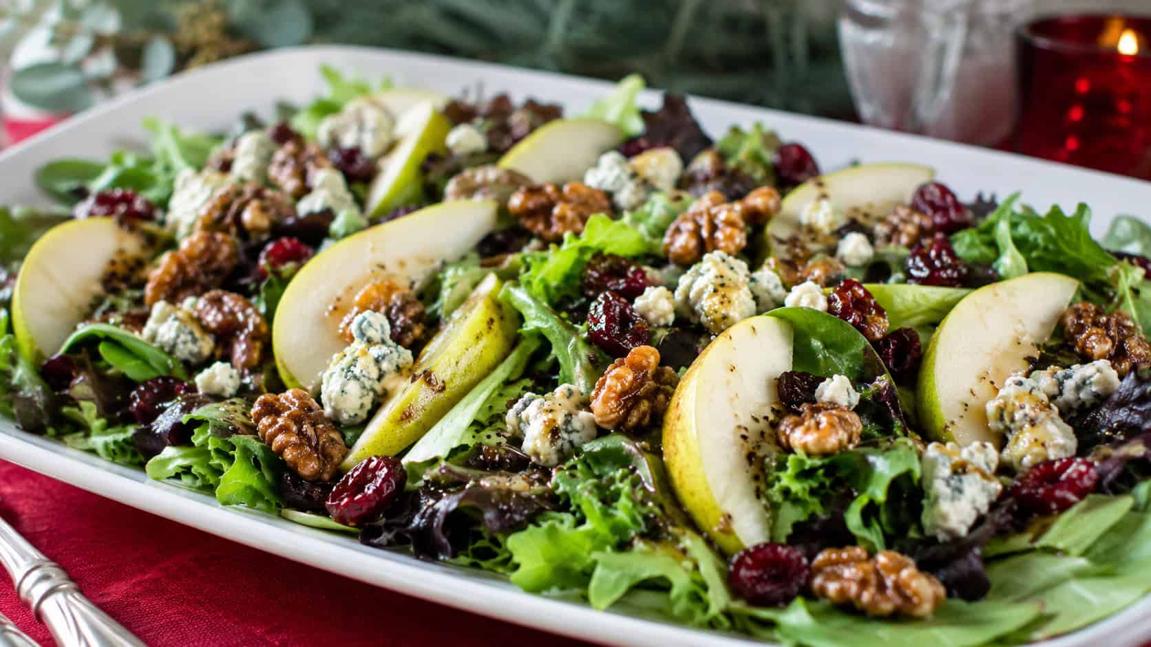 a vibrant salad with mixed greens, sliced pears, blue cheese crumbles, candied walnuts, and cranberries drizzled with vinaigrette, shown in a wide horizontal composition on a white platter over a festive table, fresh natural light, shallow depth of field, landscape orientation