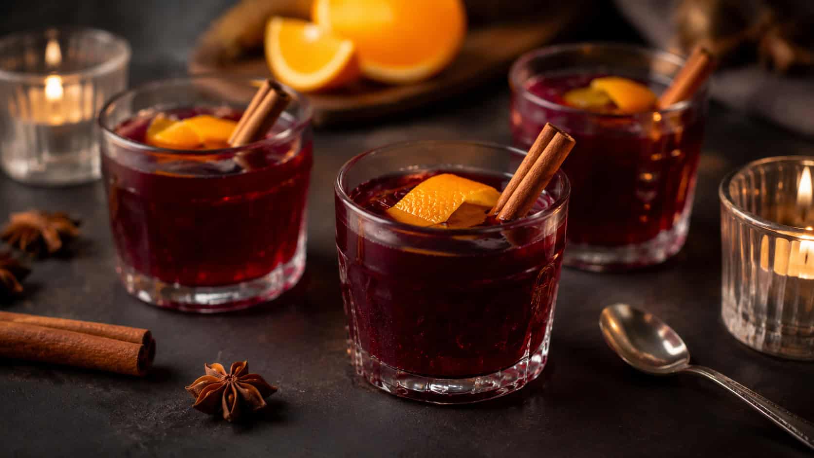 glossy deep red mulled wine jellies in small glasses garnished with orange peel and a cinnamon stick, arranged in a wide horizontal composition on a dark table with soft candlelight, warm moody light, shallow depth of field, landscape orientation