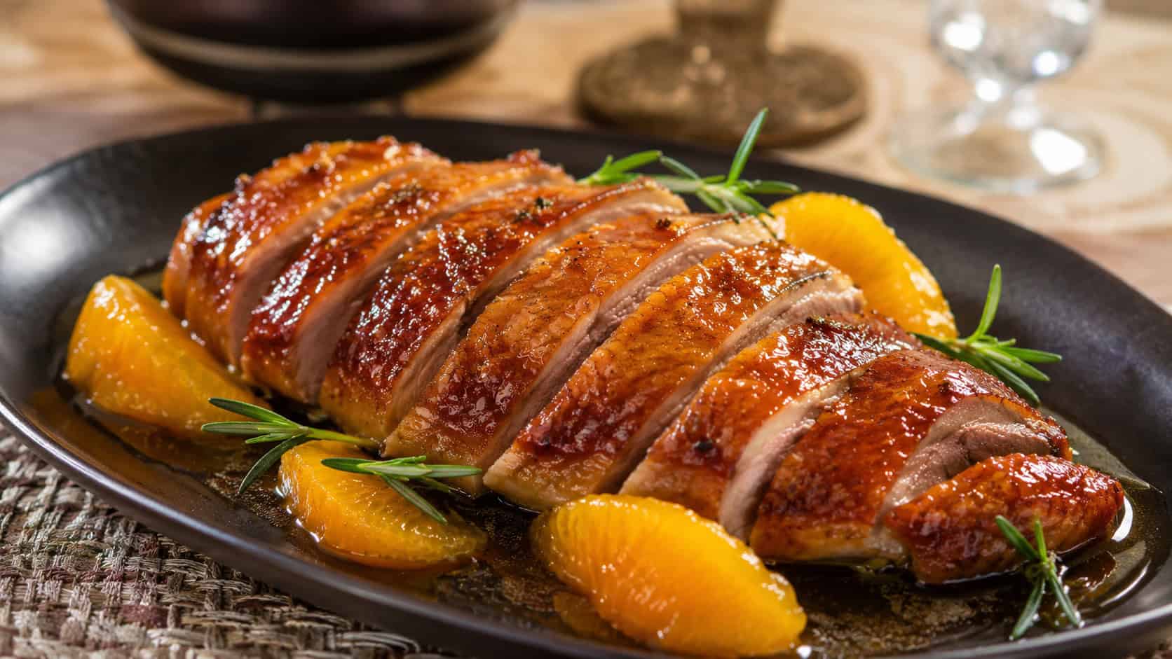 sliced roast duck breast with crispy skin glazed in orange sauce and garnished with rosemary and orange segments, shown in a wide horizontal composition on a dark plate over an elegant table, warm natural light, shallow depth of field, landscape orientation