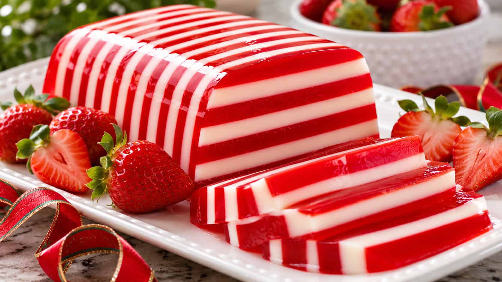 a sliced ribbon gelatin showing alternating bright red and creamy white stripes, in a wide horizontal shot plated on a white dish with fresh strawberries and a holiday ribbon nearby, bright natural light, shallow depth of field, landscape orientation