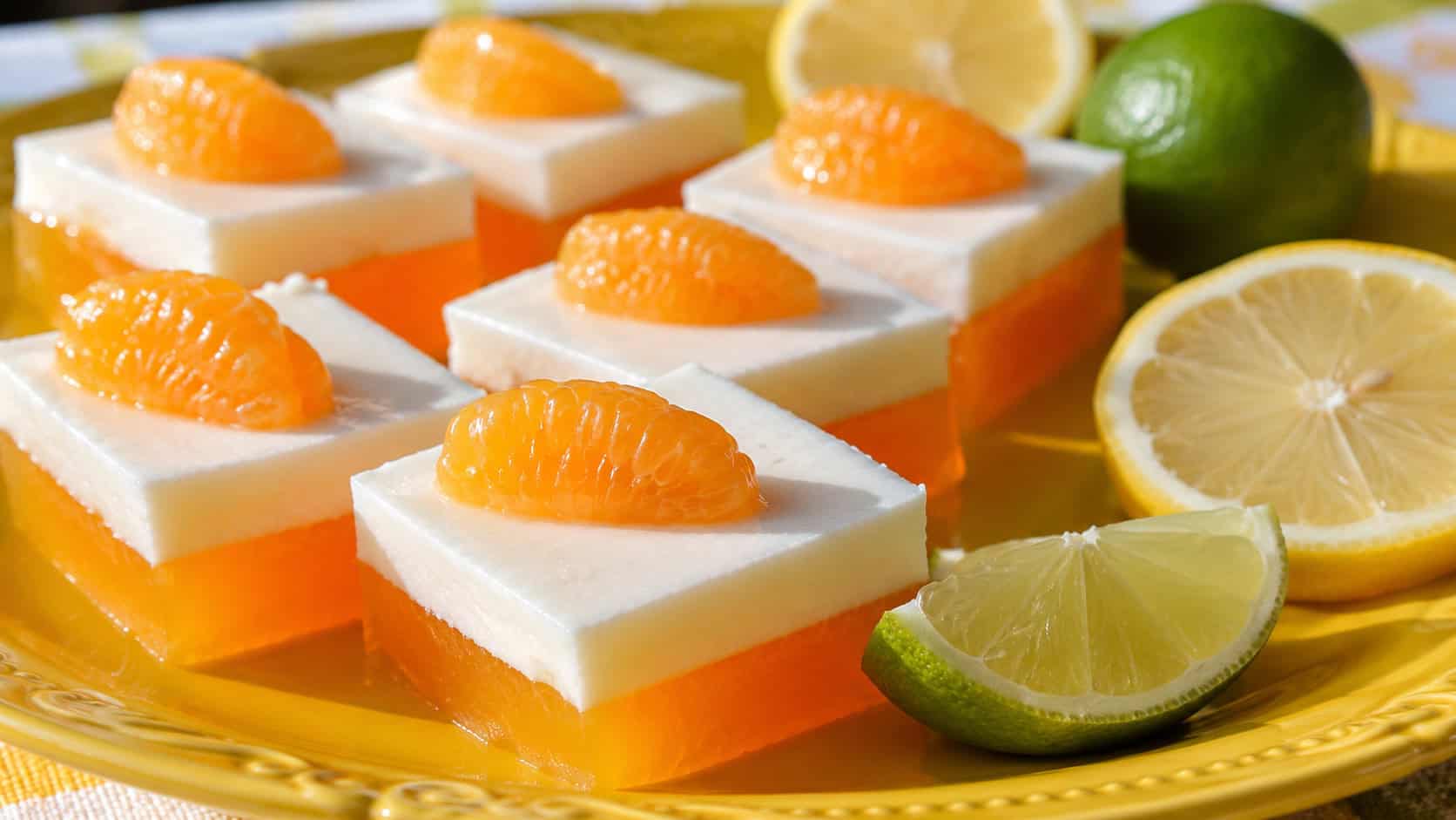 bright citrus jello squares with a creamy white top layer and mandarin orange segments, in a wide horizontal shot on a sunny yellow plate with lemon and lime slices, bright natural light, shallow depth of field, landscape orientation