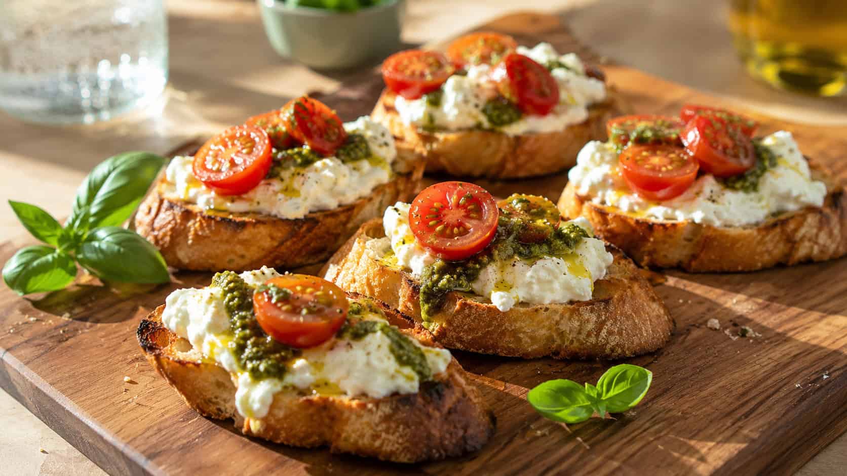 crusty toasted baguette slices topped with creamy white burrata, green pesto, and halved cherry tomatoes, shown in a wide horizontal shot on a wooden board with fresh basil leaves, on a sunny table, bright natural light, shallow depth of field, landscape orientation