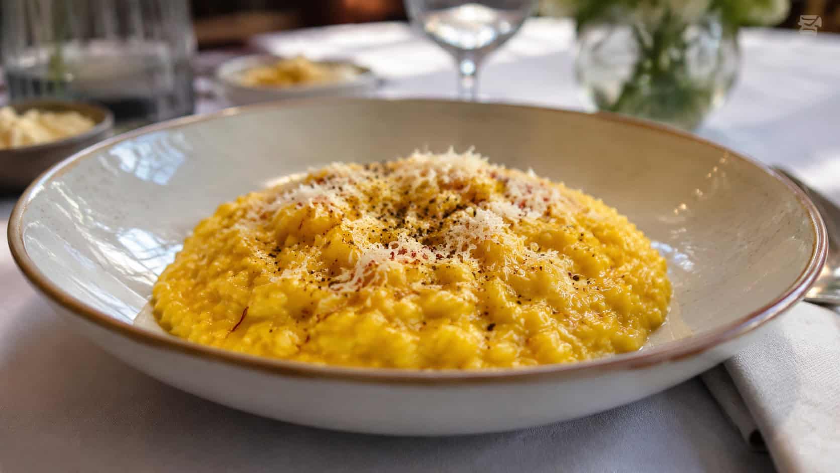 a creamy golden saffron risotto with a glossy sheen and a dusting of Parmigiano, shown in a wide horizontal shot in a shallow bowl with a sprinkle of black pepper, on an elegant table, soft natural light, shallow depth of field, landscape orientation
