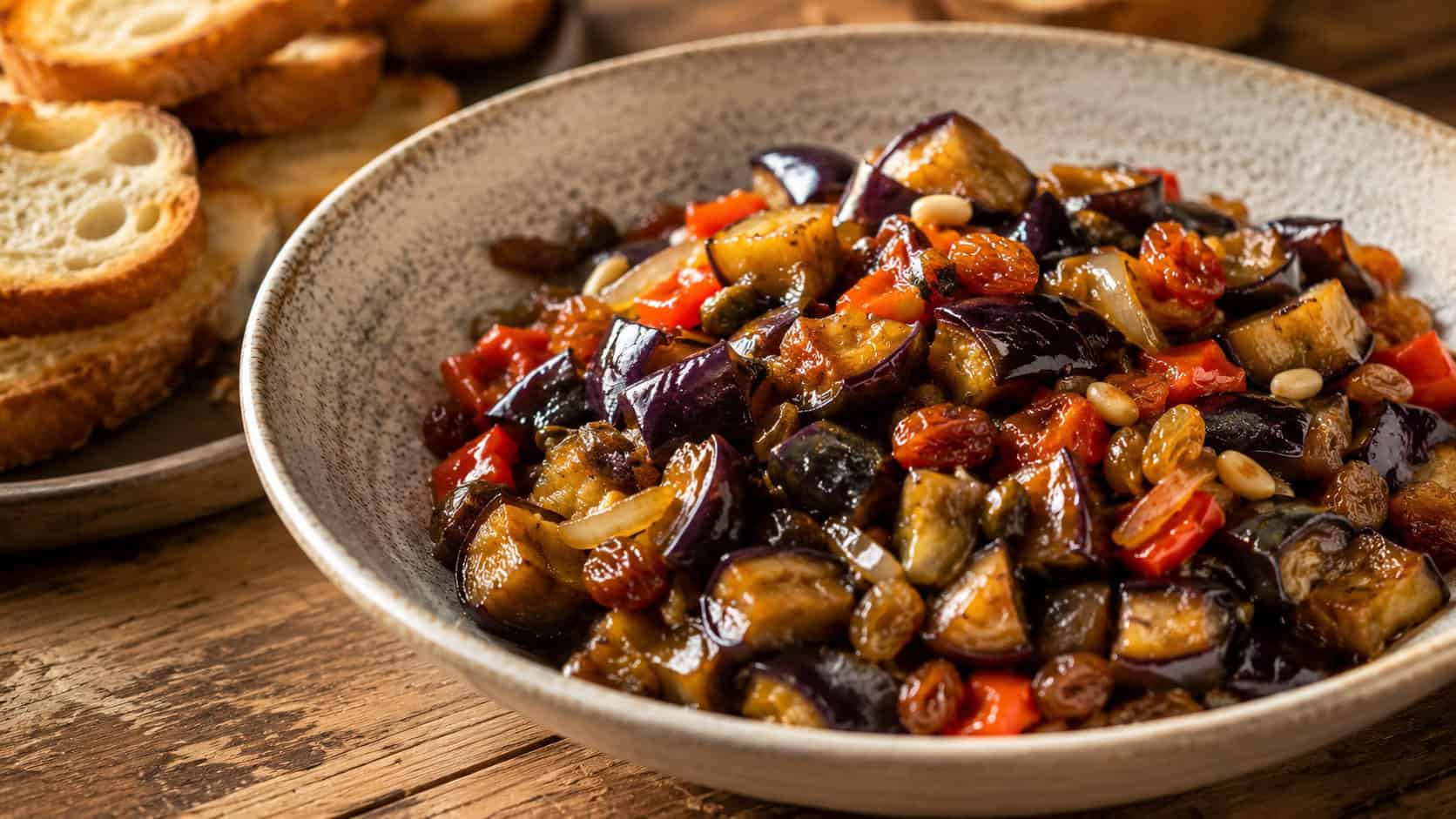 a glossy sweet and sour eggplant caponata flecked with pine nuts and raisins, shown in a wide horizontal shot in a rustic bowl with crostini alongside, on a wooden table, warm natural light, shallow depth of field, landscape orientation