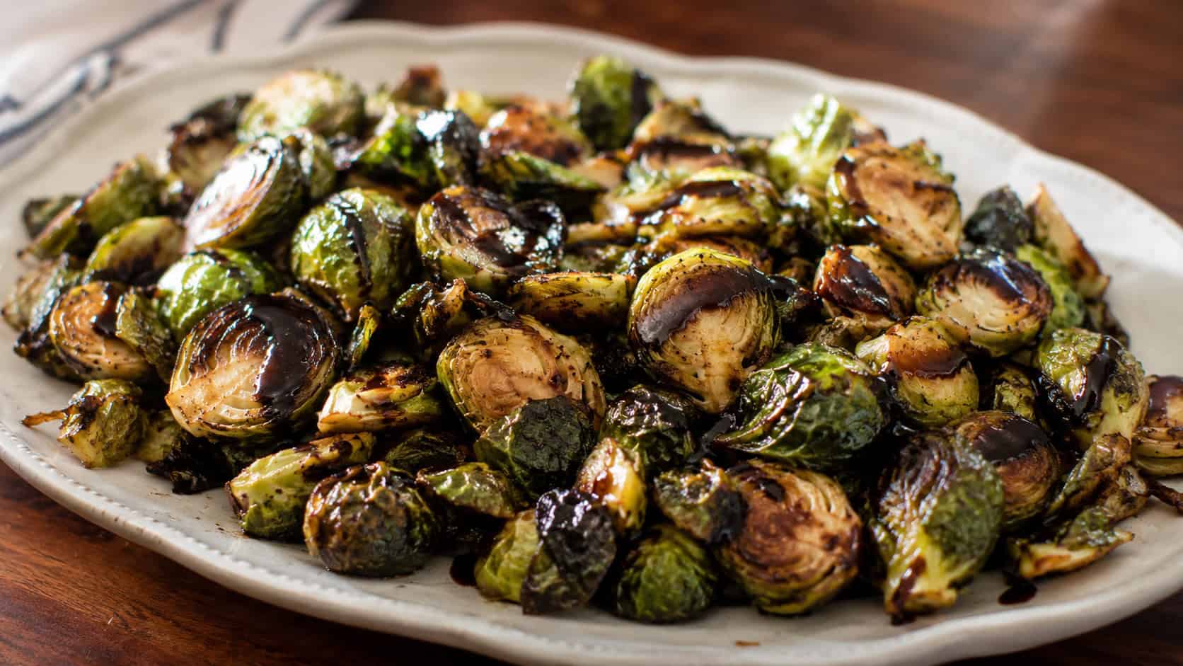 crispy roasted Brussels sprouts drizzled with dark balsamic glaze spread wide across a serving platter, charred edges visible, warm natural light, shallow depth of field, horizontal landscape composition