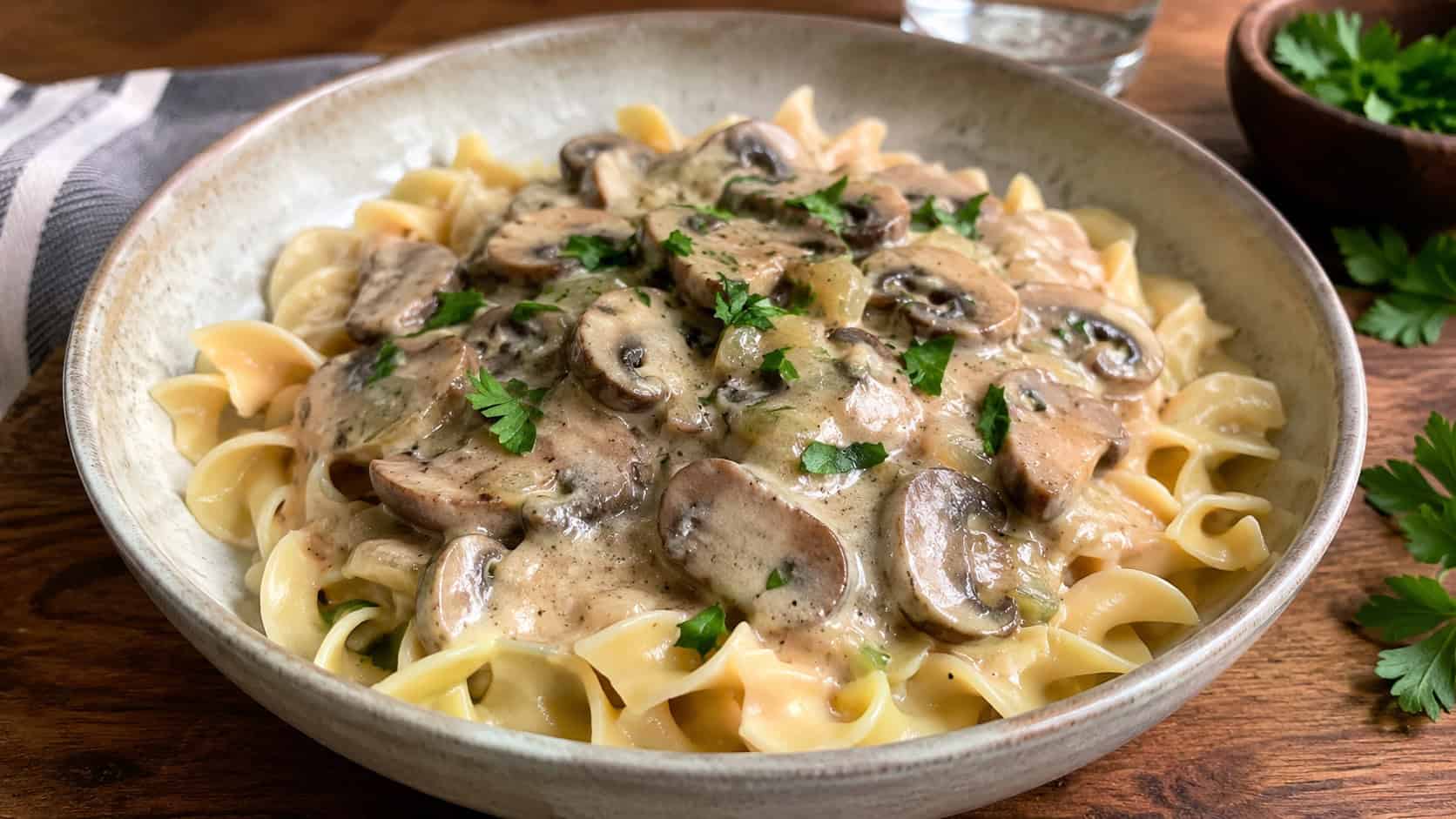 a bowl of creamy mushroom stroganoff over wide noodles shown wide across a wooden table, fresh parsley sprinkled on top, soft natural light, shallow depth of field, horizontal landscape composition