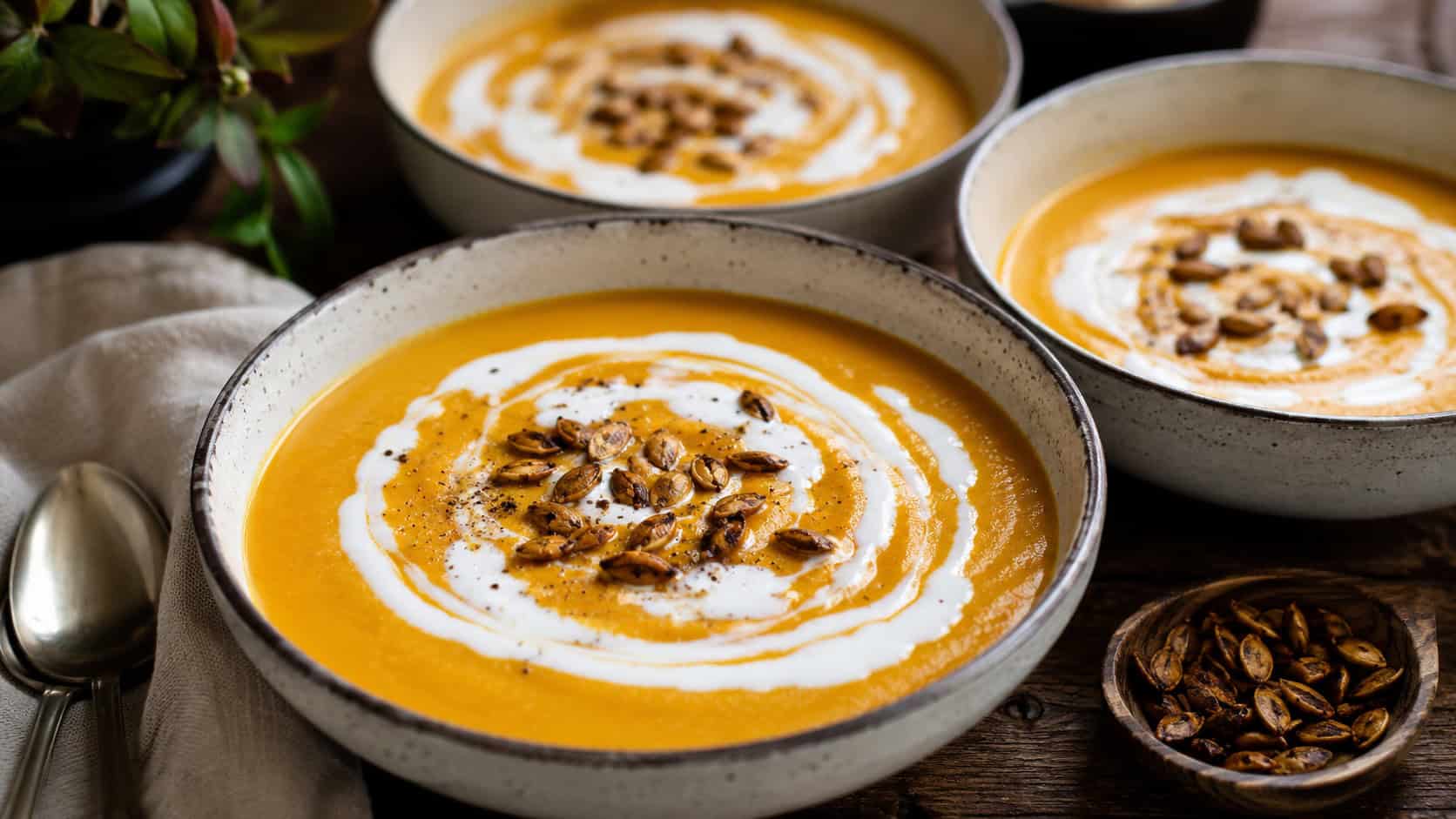 bowls of velvety orange pumpkin soup swirled with coconut cream shown wide across a wooden table, toasted seeds sprinkled on top, soft natural light, shallow depth of field, landscape orientation