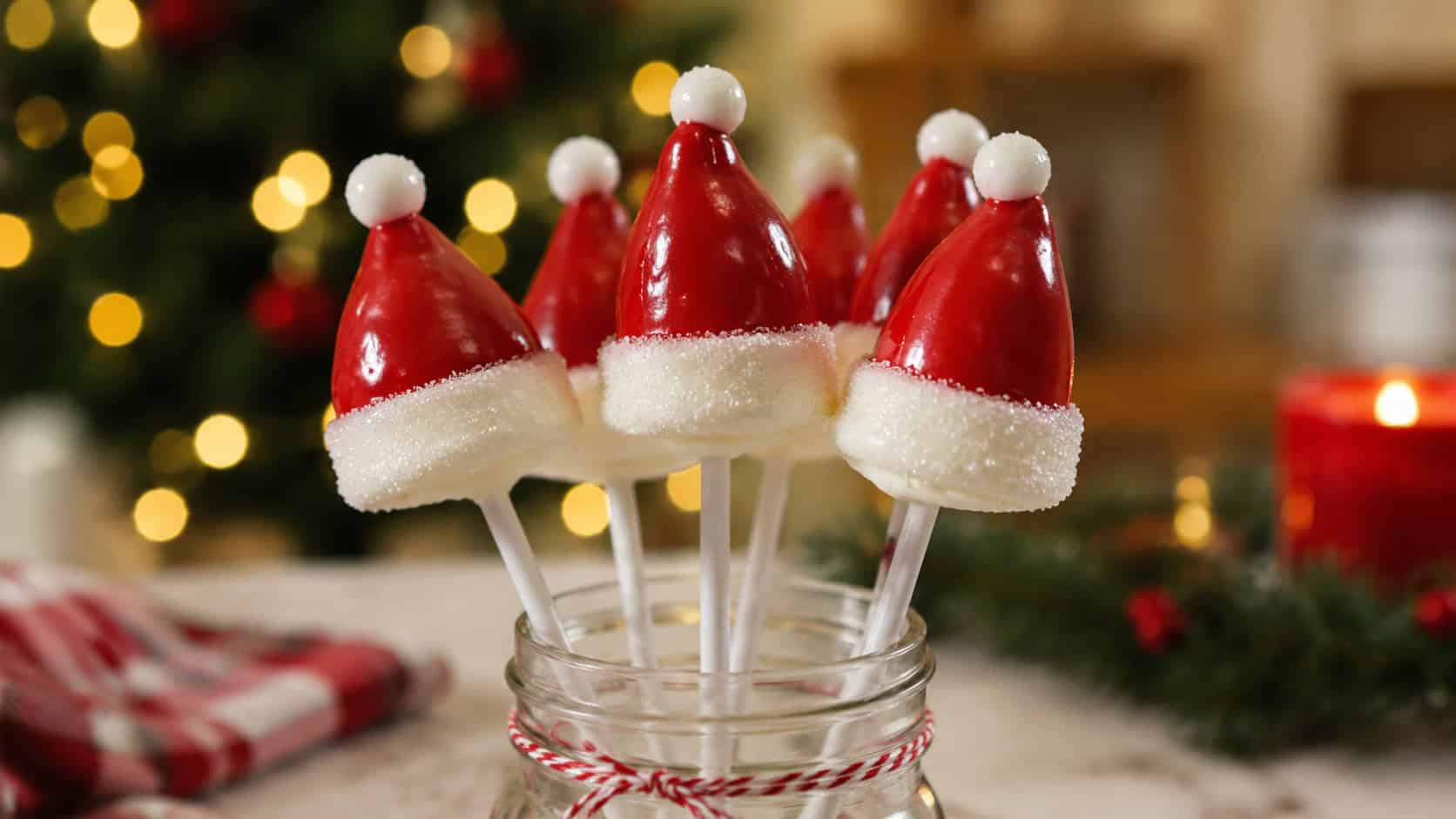 a cluster of Santa-hat marshmallow pops with glossy red coating and white sugared brims standing upright, in a wide horizontal shot in a mason jar on a festive table with twinkling lights blurred behind, warm natural light, shallow depth of field, landscape orientation