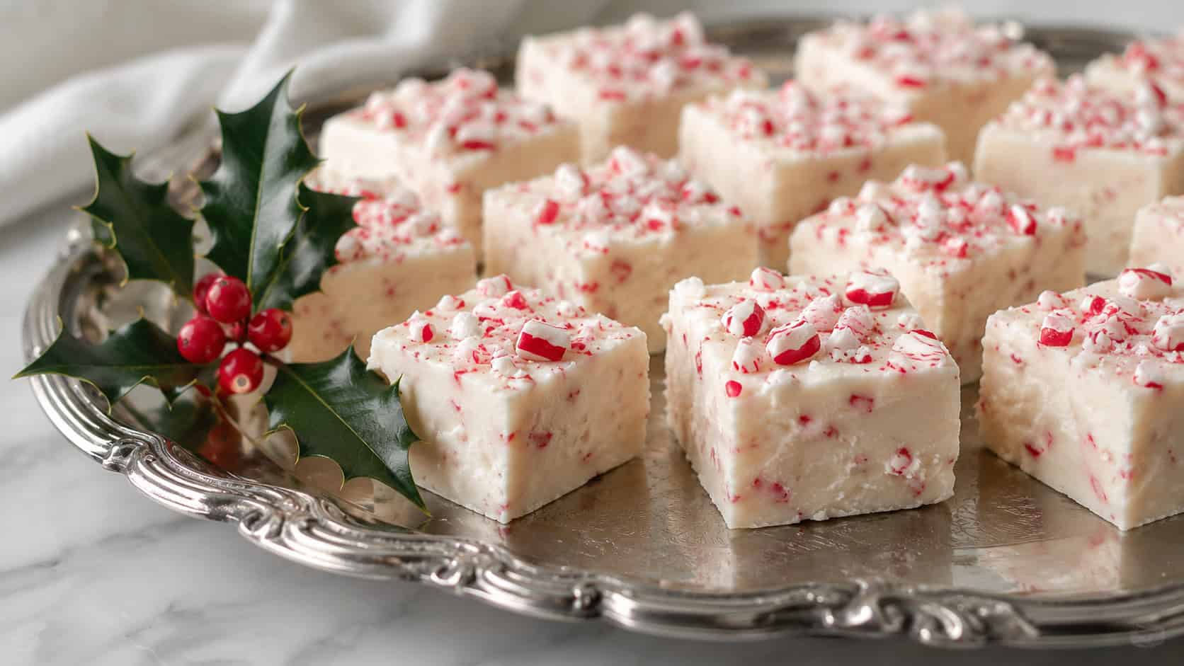neat squares of creamy white peppermint marshmallow fudge topped with crushed red and white candy canes, in a wide horizontal shot arranged on a vintage silver tray with a sprig of holly, soft natural light, shallow depth of field, landscape orientation