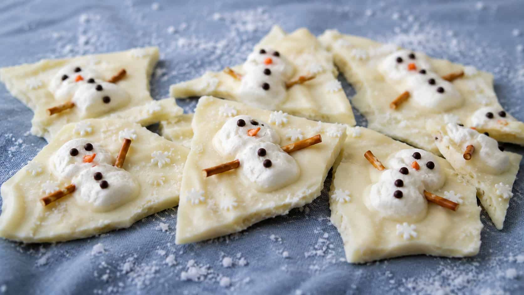 a sheet of white chocolate bark broken into pieces showing slumped marshmallow snowmen with candy faces and pretzel arms, in a wide horizontal shot on a blue linen napkin dusted with powdered sugar, cool natural light, shallow depth of field, landscape orientation