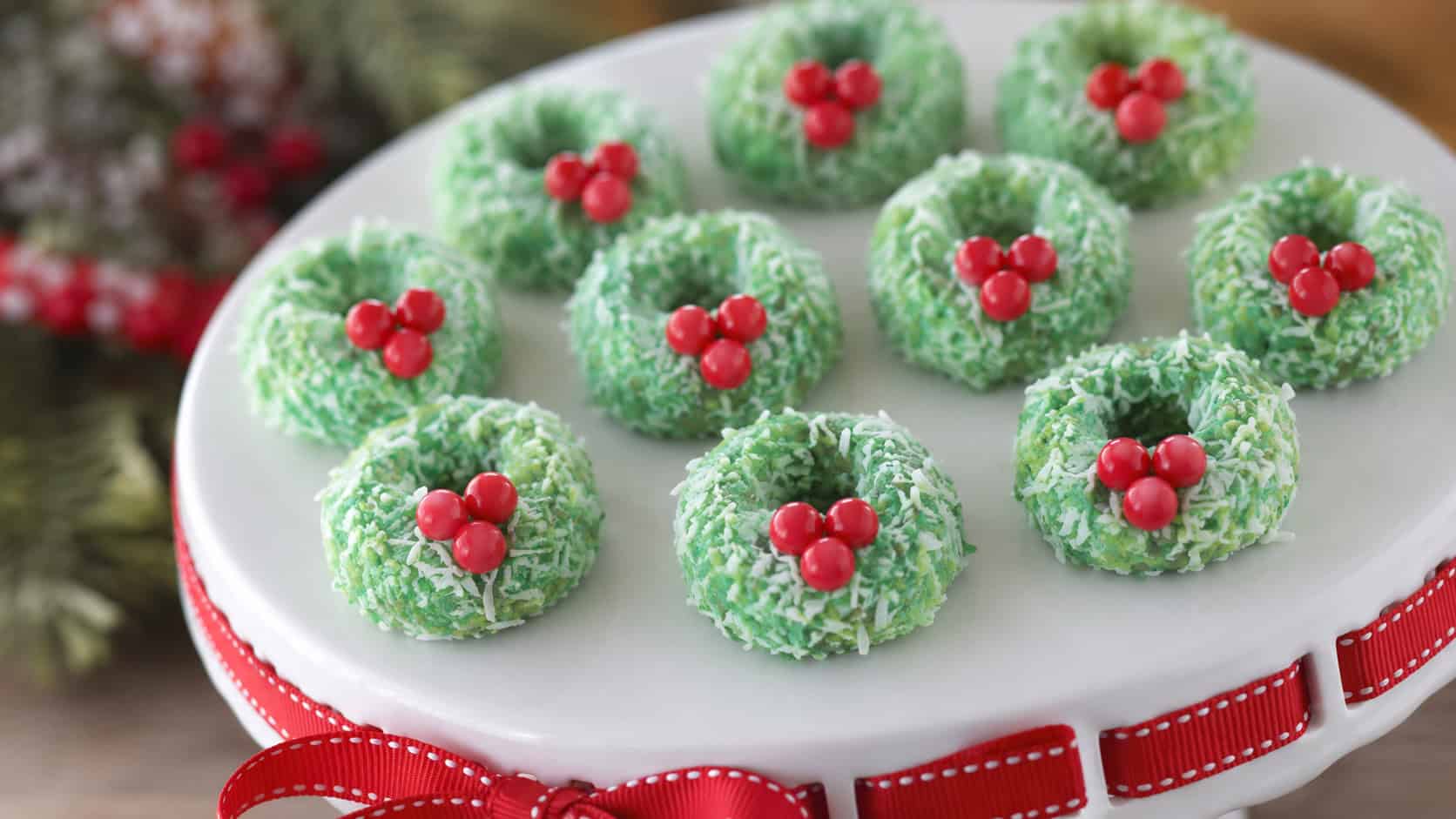 edible mini wreaths made of green coconut-coated marshmallows with red candy berries, in a wide horizontal shot arranged on a white cake stand with a red ribbon, soft natural light, shallow depth of field, landscape orientation