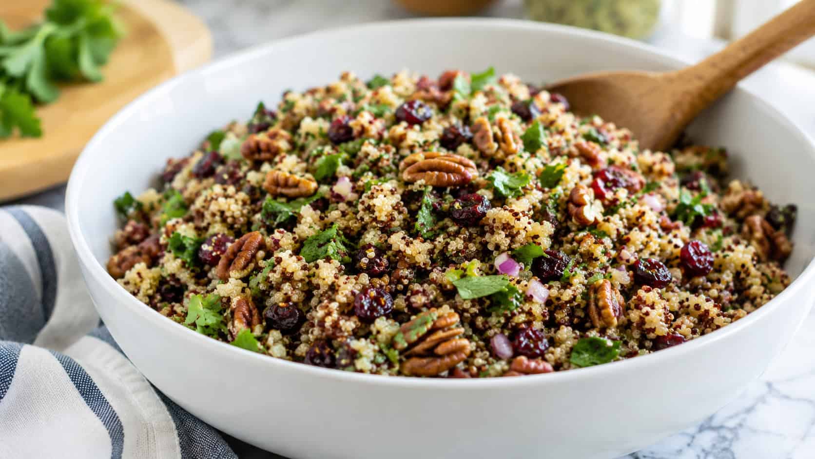 a colorful quinoa salad studded with red cranberries, toasted pecans, and green parsley, shown in a wide horizontal shot in a large white serving bowl with a wooden spoon, on a bright kitchen counter, fresh natural light, shallow depth of field, landscape orientation