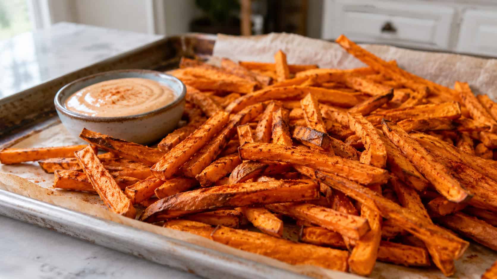 a heap of crispy golden-orange sweet potato fries dusted with paprika, shown in a wide horizontal shot piled on a parchment-lined sheet pan with a small bowl of dip, on a kitchen counter, bright natural light, shallow depth of field, landscape orientation