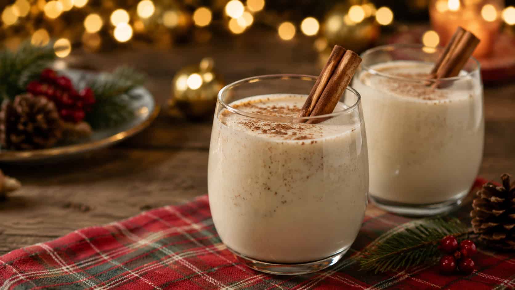 two glasses of creamy pale eggnog dusted with nutmeg and garnished with a cinnamon stick, shown in a wide horizontal shot on a festive table with twinkling lights softly blurred behind, cozy warm light, shallow depth of field, landscape orientation