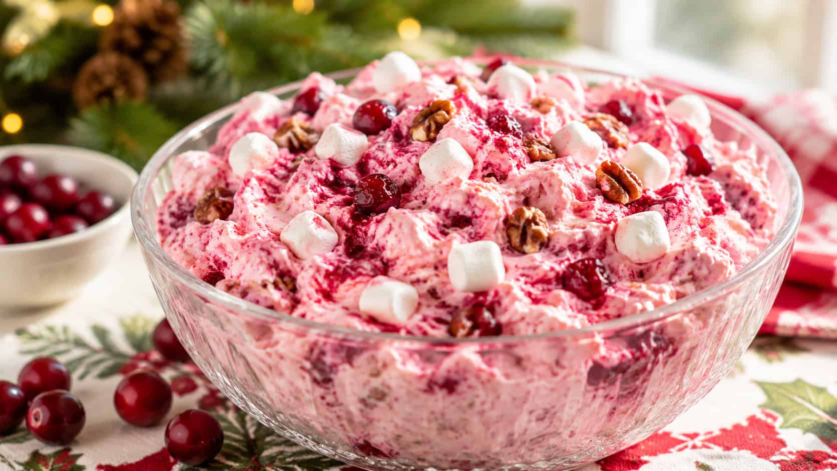 a fluffy pink cranberry cream cheese fluff swirled with red and dotted with marshmallows and pecans, in a wide horizontal shot in a clear glass bowl on a holiday table with fresh cranberries scattered nearby, bright natural light, shallow depth of field, landscape orientation
