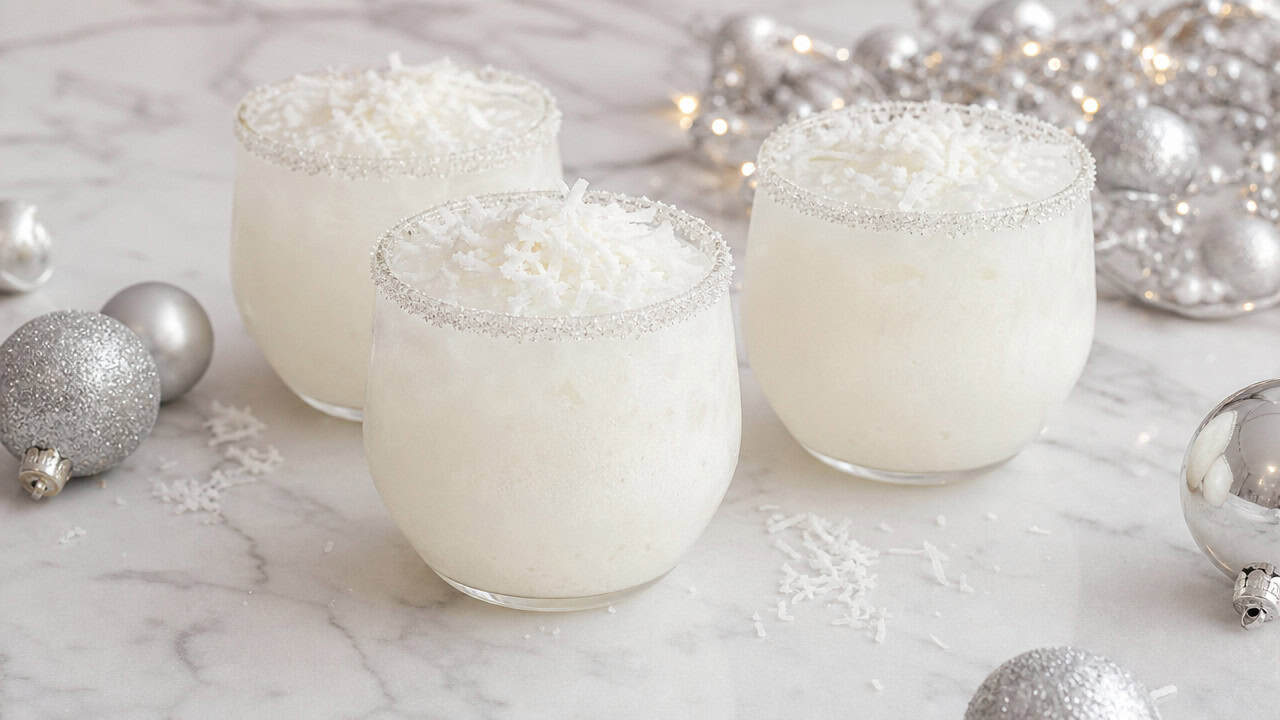 creamy white Christmas margaritas with sparkling sugar rims and shredded coconut arranged in a wide horizontal shot on a white marble surface, silver ornaments and soft light nearby, shallow depth of field, landscape orientation