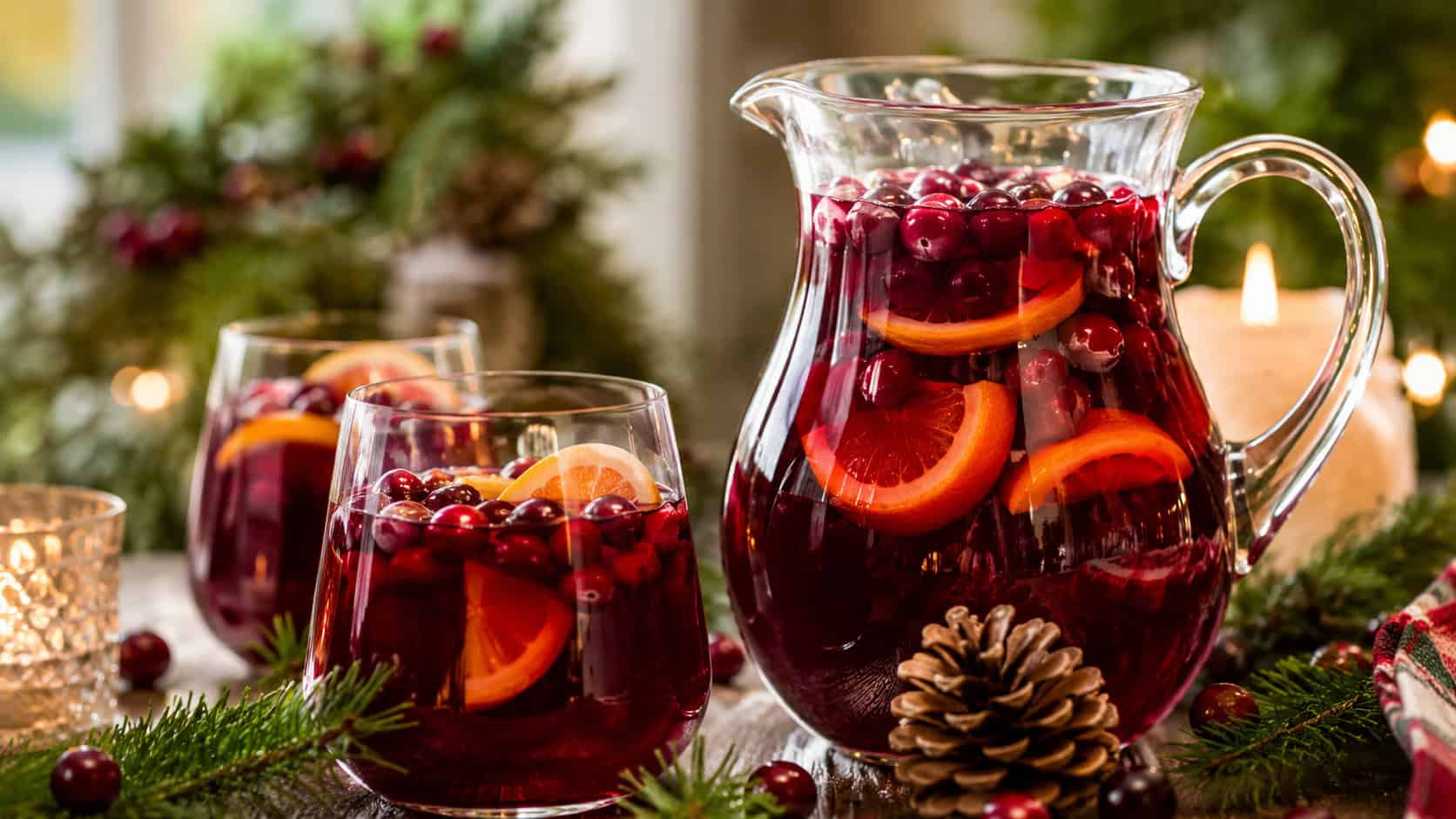 a pitcher and glasses of red cranberry sangria full of floating cranberries and orange slices arranged in a wide horizontal shot on a festive table, pine and candles nearby, warm natural light, shallow depth of field, landscape orientation