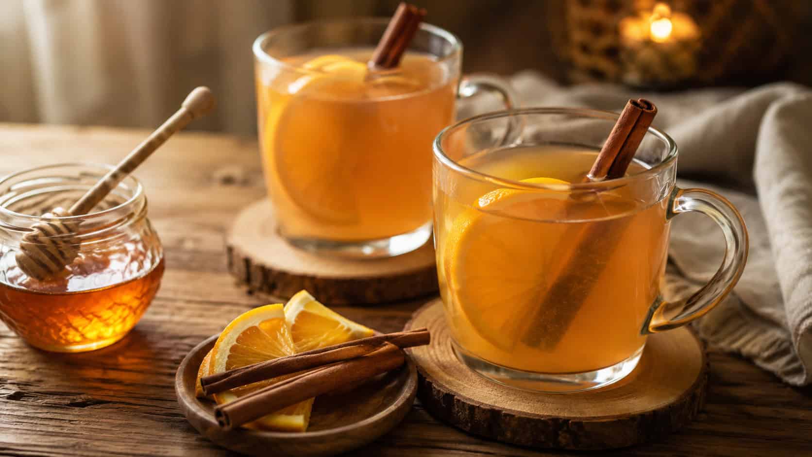 warm orange brandy hot toddies in glass mugs with orange slices and cinnamon sticks arranged in a wide horizontal shot on a wooden table, honey jar and soft light nearby, warm cozy light, shallow depth of field, landscape orientation