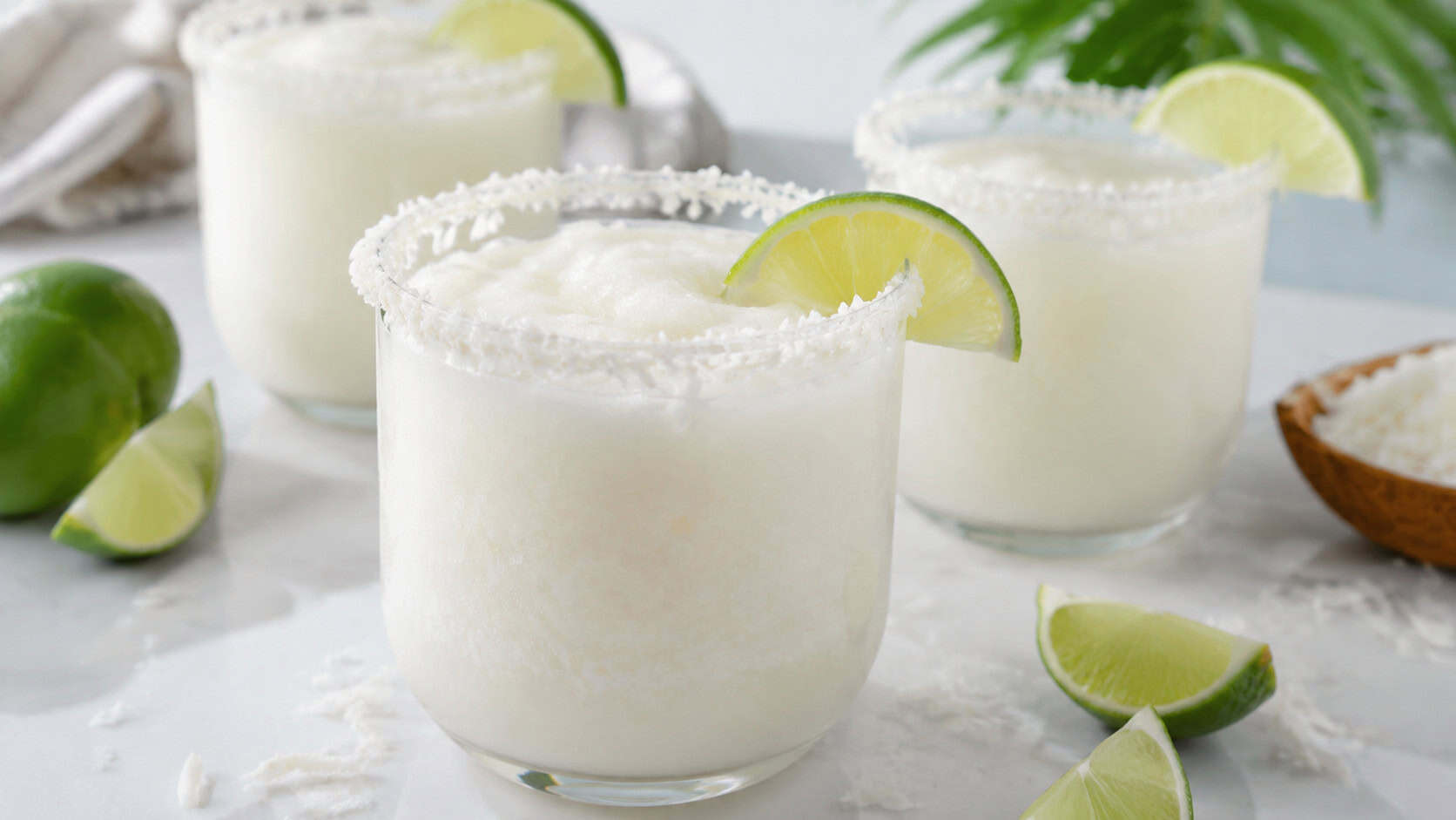 frosty blended coconut margaritas with shredded coconut rims arranged in a wide horizontal shot on a bright white surface, lime wedges and soft daylight nearby, shallow depth of field, landscape orientation