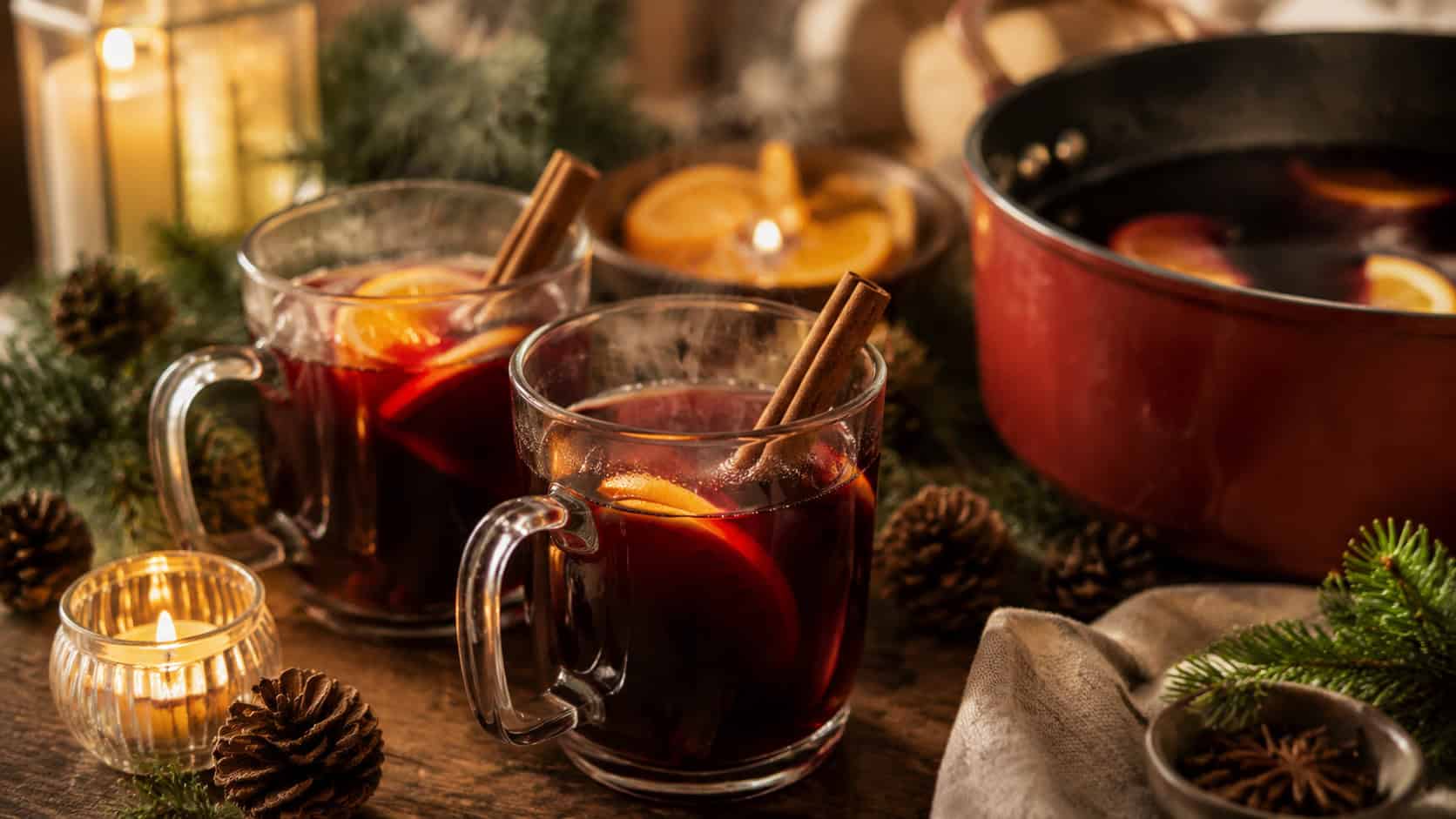 steaming mulled wine in glass mugs with cinnamon sticks and orange slices arranged in a wide horizontal shot beside a simmering pot, pine and candles on a rustic table, warm cozy light, shallow depth of field, landscape orientation