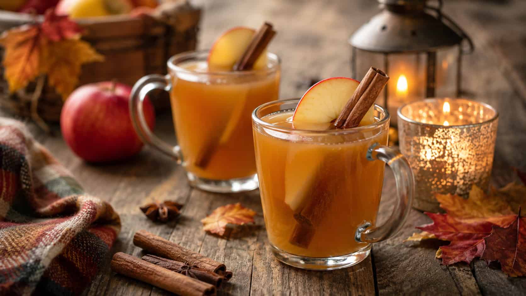 warm spiced apple cider with bourbon in glass mugs garnished with apple slices and cinnamon sticks arranged in a wide horizontal shot on a rustic wooden table, autumn leaves and candles nearby, warm natural light, shallow depth of field, landscape orientation