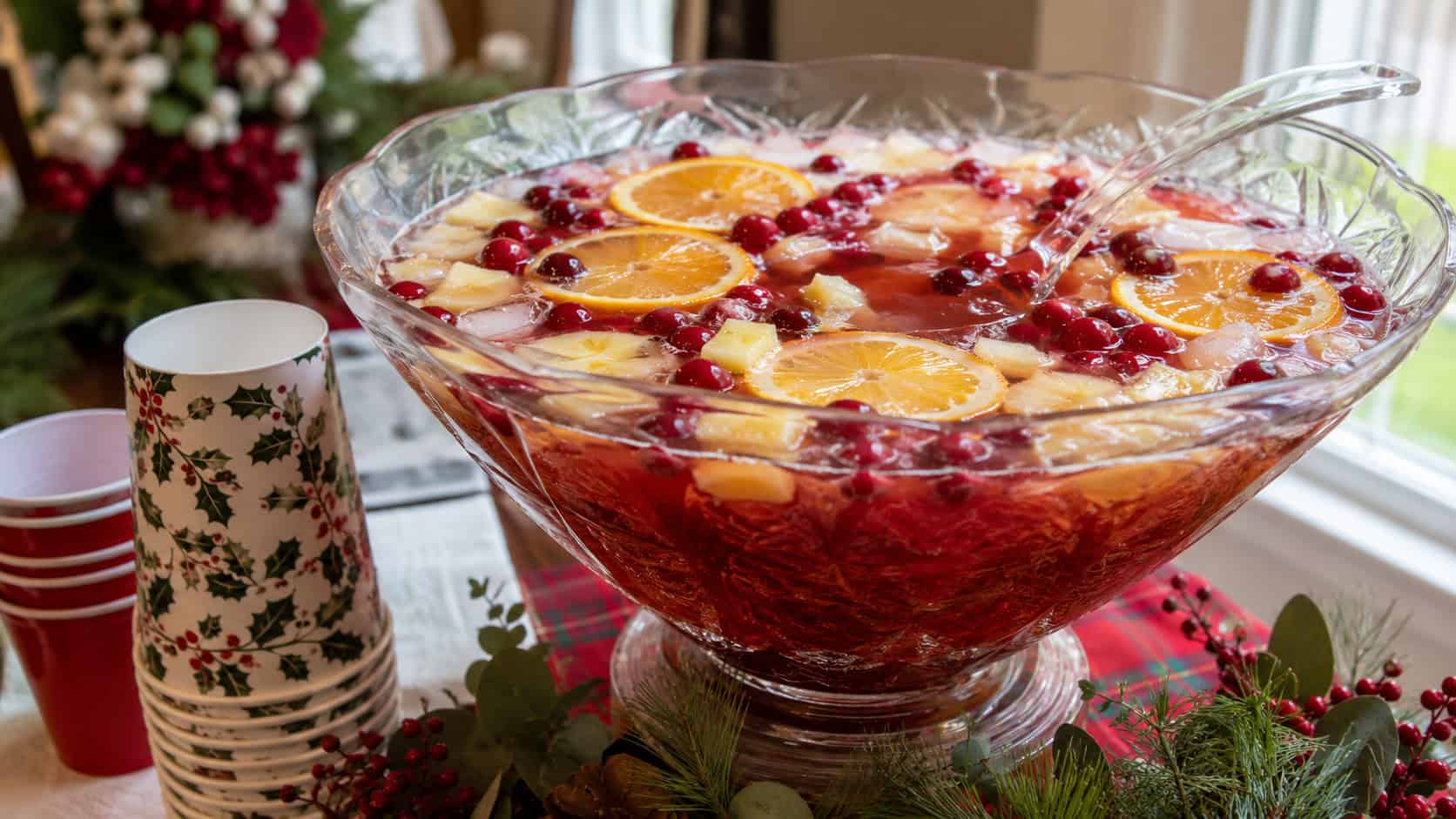 a large glass punch bowl of pineapple cranberry holiday punch with floating orange slices and cranberries arranged in a wide horizontal shot on a party table, festive cups and greenery nearby, bright natural light, shallow depth of field, landscape orientation