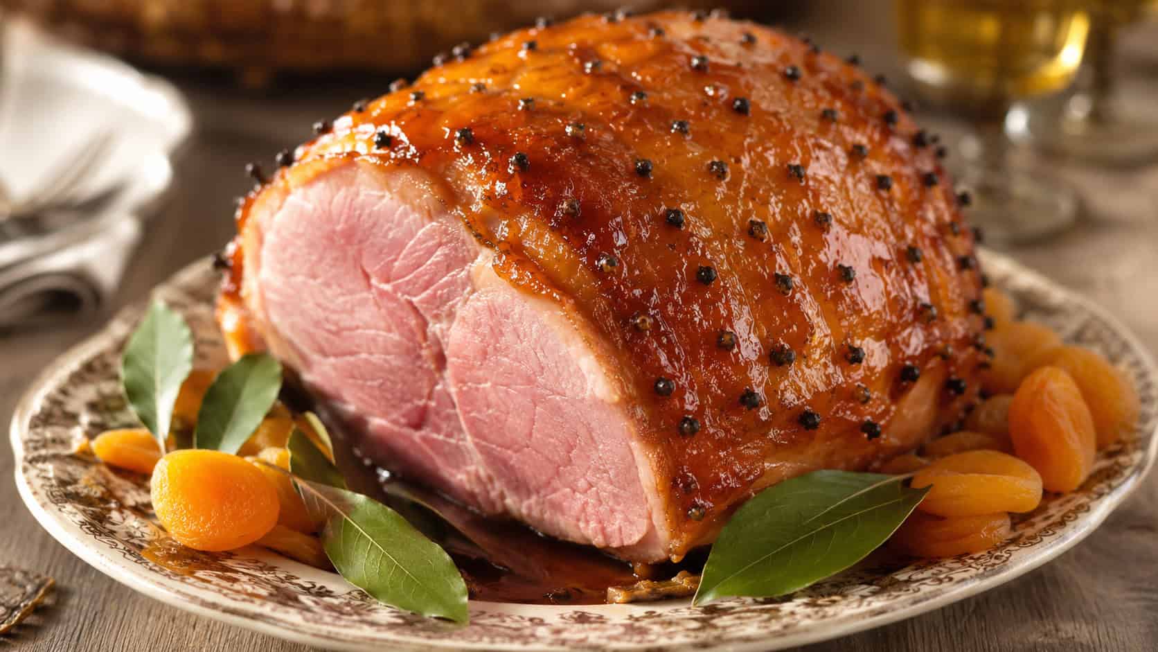 a clove-studded ham with a golden apricot glaze on a vintage platter in a wide horizontal shot, glossy amber surface, dried apricots and bay leaves alongside, warm natural light, shallow depth of field, landscape orientation