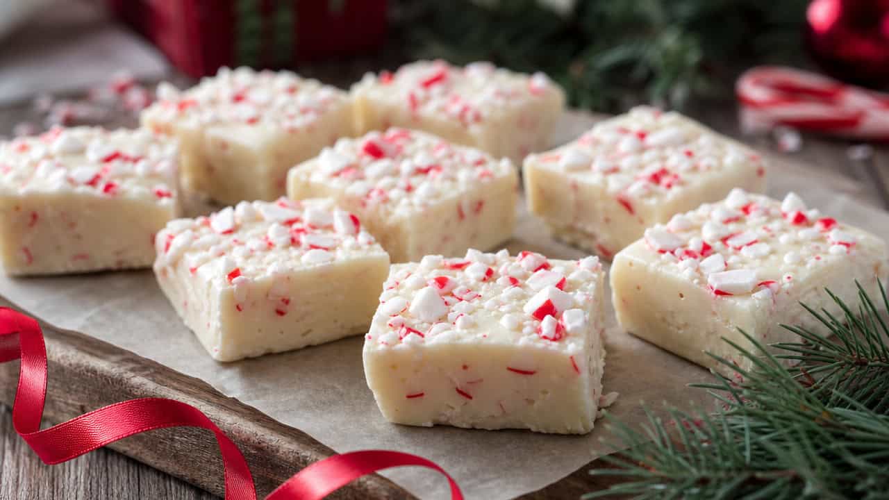 squares of white peppermint bark fudge topped with crushed candy cane arranged in a wide horizontal shot on parchment over a wooden board, red ribbon and pine nearby, soft natural light, shallow depth of field, landscape orientation
