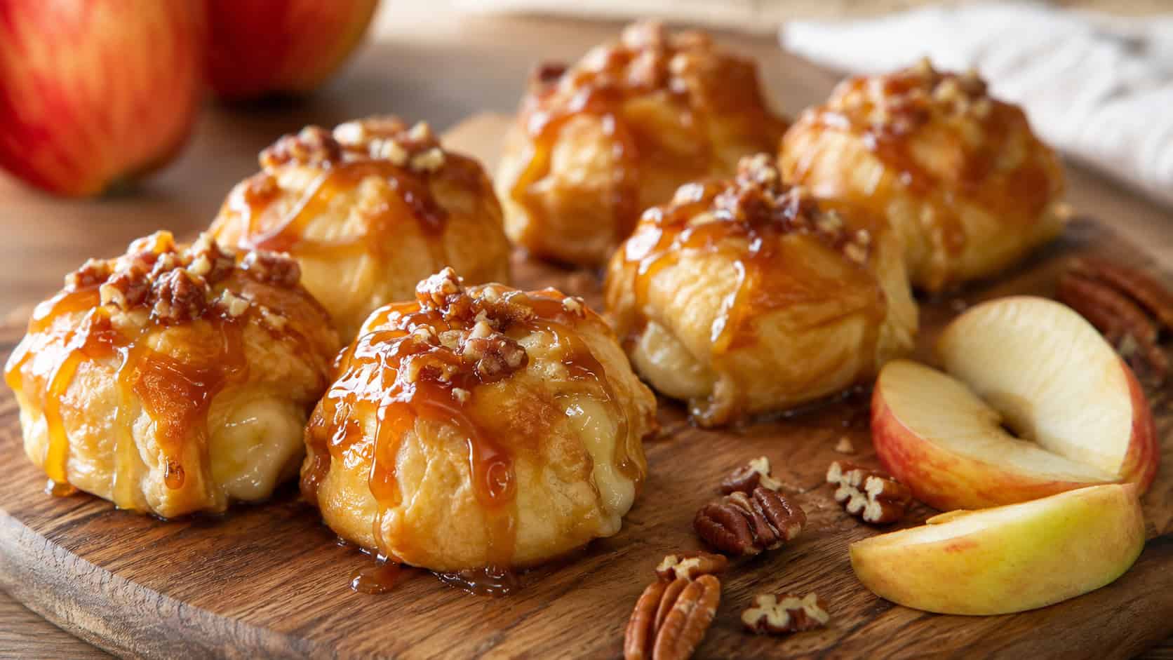 golden caramel apple brie pastry bites drizzled with caramel arranged in a wide horizontal shot on a wooden serving board, apple slices and pecans scattered nearby, warm natural light, shallow depth of field, landscape orientation