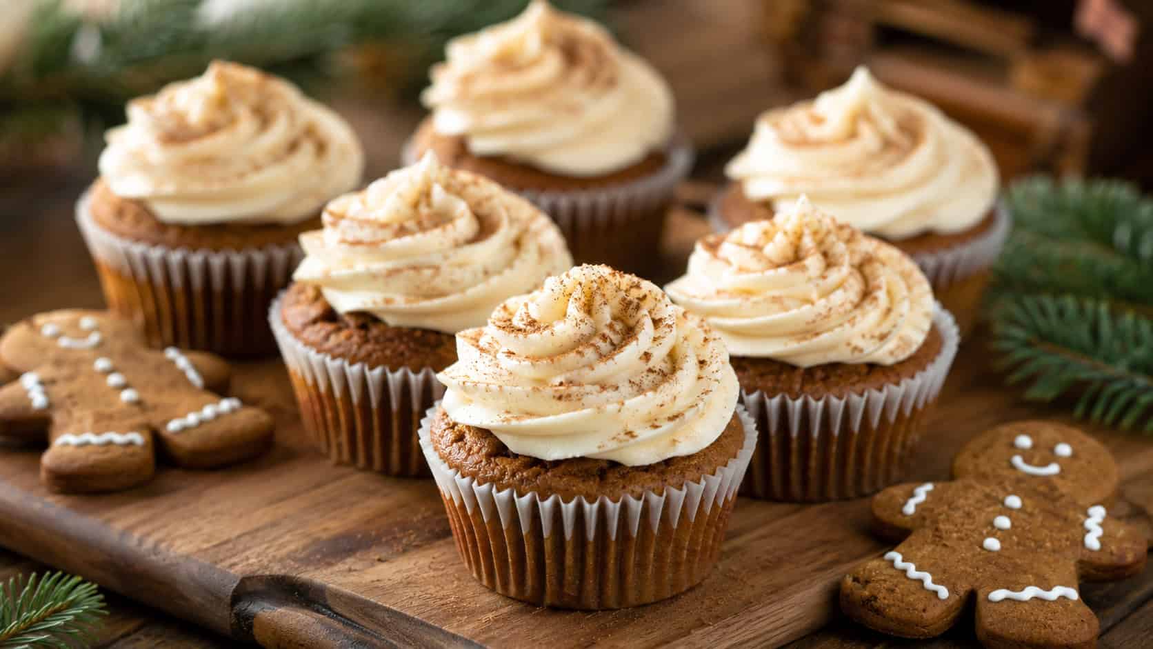gingerbread cupcakes with swirled cream cheese frosting and a dusting of cinnamon arranged in a wide horizontal shot on a wooden board, gingerbread men and pine sprigs nearby, warm natural light, shallow depth of field, landscape orientation