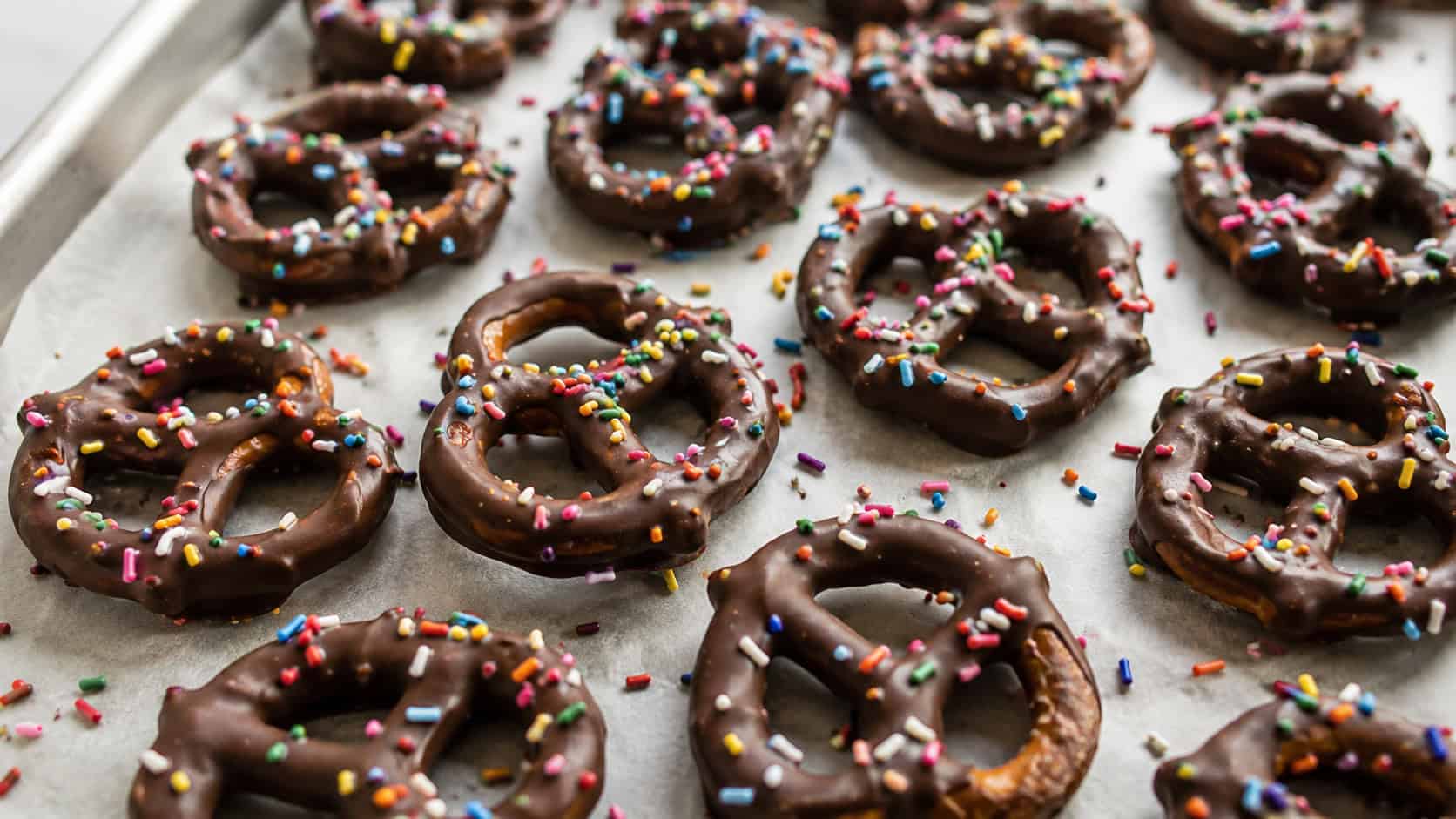 chocolate-dipped pretzel twists topped with colorful sprinkles arranged wide across a parchment-lined tray, scattered sprinkles around, bright natural light, shallow depth of field, horizontal landscape composition