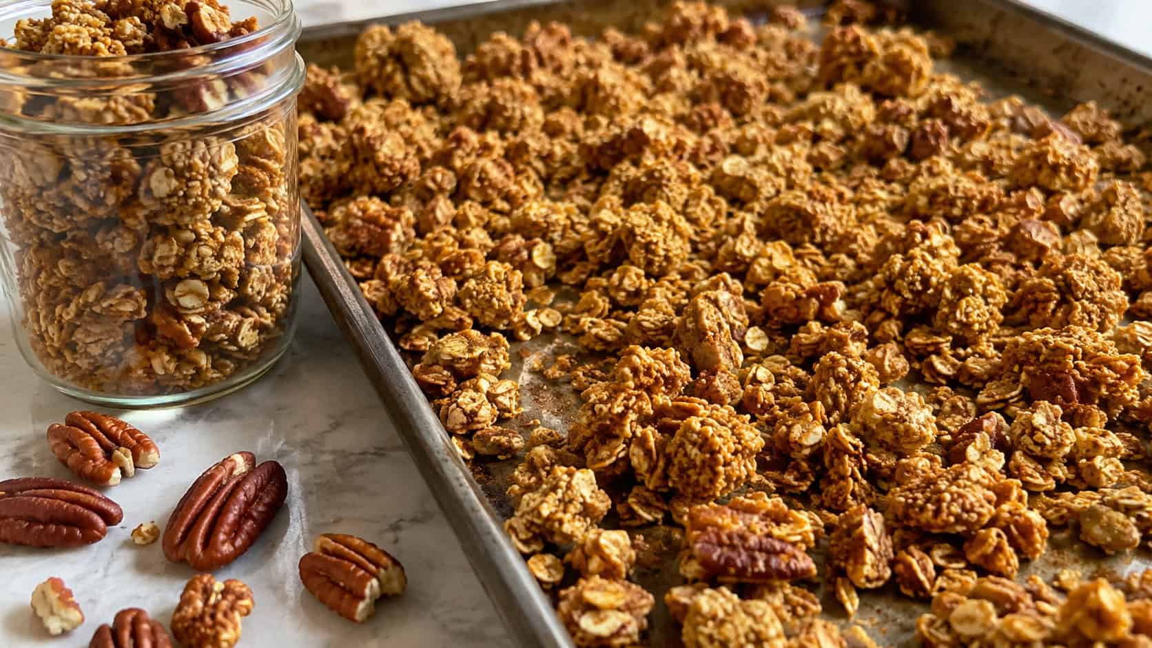 golden pumpkin spice granola clusters spread wide across a baking tray, a filled jar and scattered pecans nearby, warm natural light, shallow depth of field, landscape orientation