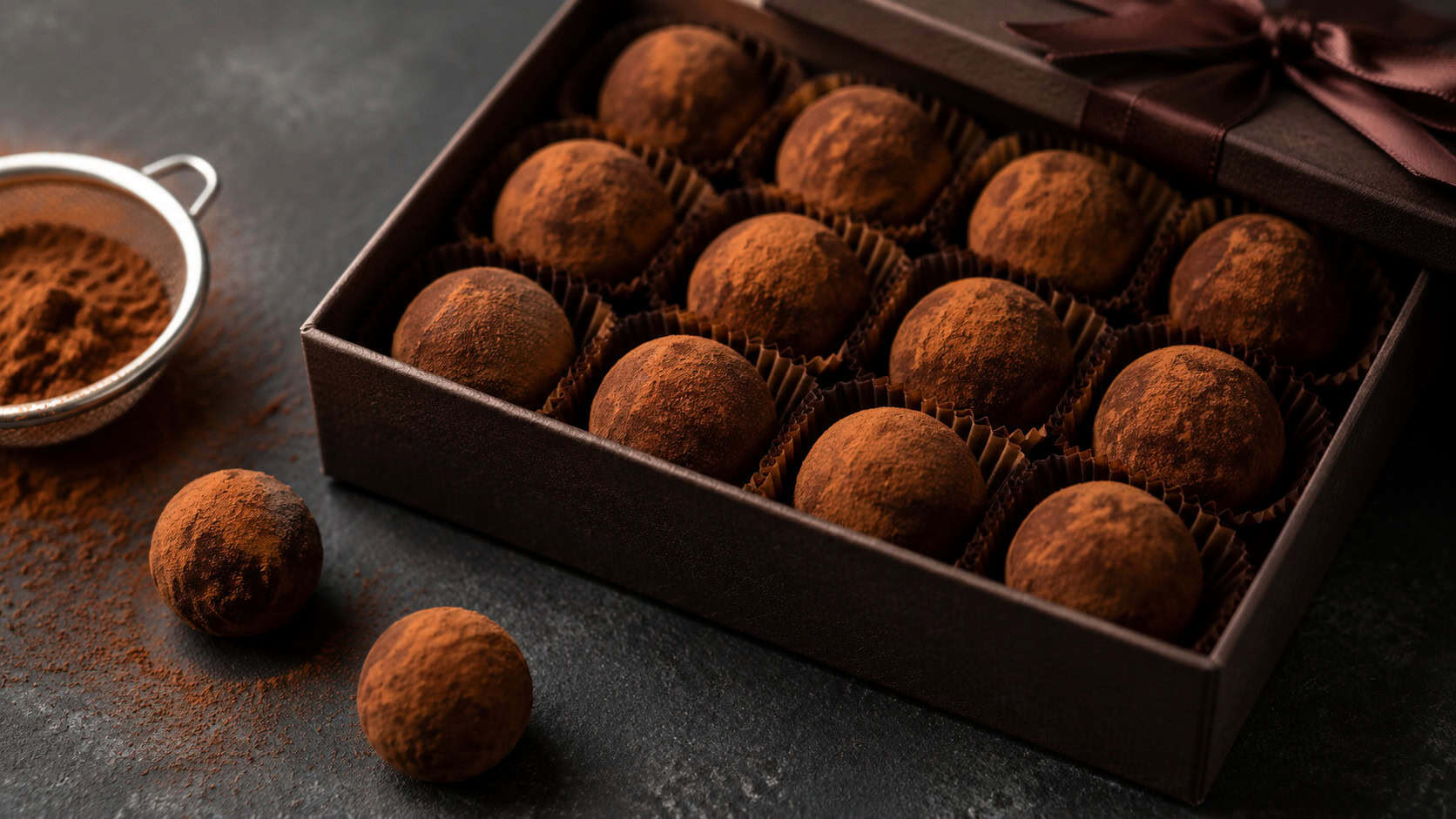 cocoa-dusted dark chocolate truffles nestled in a gift box shown wide across a dark surface, a few loose truffles and cocoa nearby, moody natural light, shallow depth of field, horizontal landscape composition