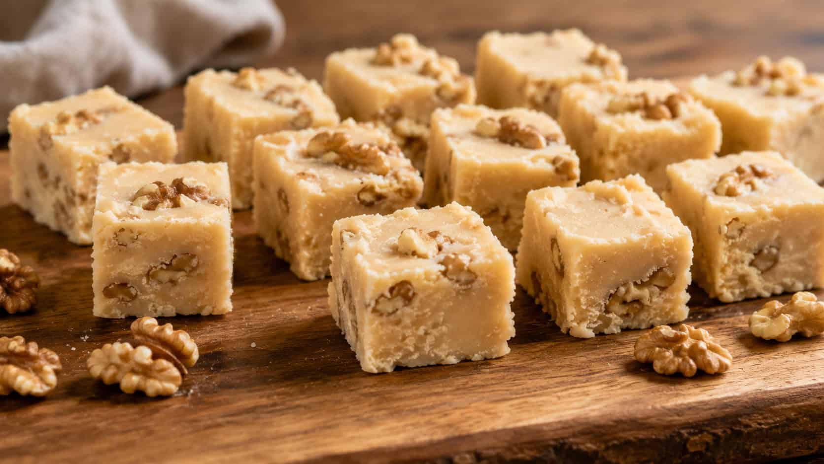squares of pale maple fudge studded with walnut pieces arranged wide on a wooden board, a few toasted walnuts scattered nearby, warm natural light, shallow depth of field, horizontal landscape composition